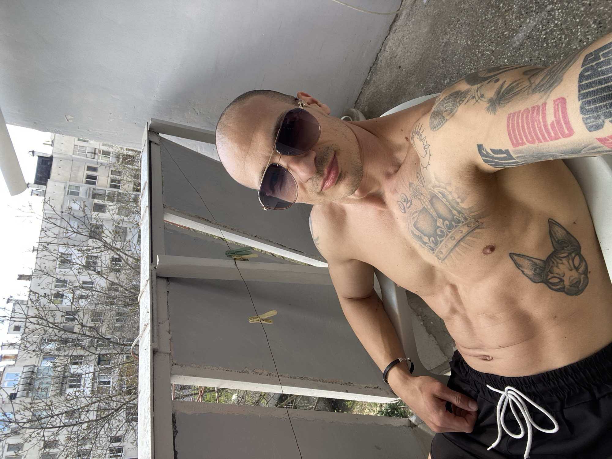 A shirtless man with tattoos and sunglasses poses on a balcony in Pleven, Bulgaria. The man has a shaved head and is wearing dark sunglasses. He is tattooed on his upper body. He is wearing black shorts with a white drawstring. The balcony has a metal railing with clothespins attached to a line. Behind the railing, there is a view of buildings and trees. The sky is overcast.