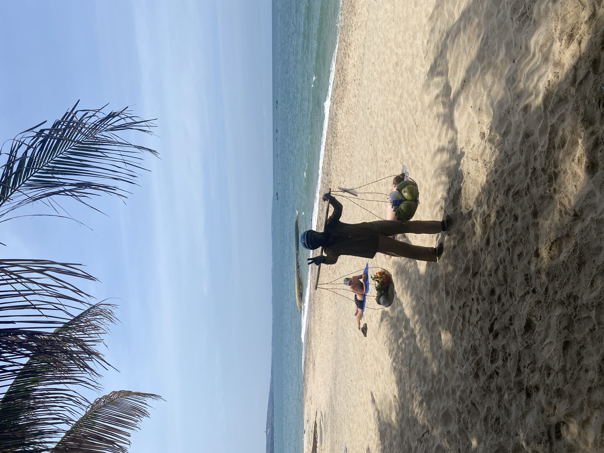 In Phu Thuy Ward, Vietnam, on a sunny beach, a person walks with a yoke across their shoulders, balancing baskets of fruit. The left basket contains coconuts, while the right basket holds a mixture of fruits including coconuts, oranges, and possibly other produce. The person is wearing a dark shirt, khaki pants, and a dark helmet. They are raising their right hand in a peace sign gesture towards the viewer. In the background, another person is lying on a blue mat on the sand, sunbathing. The ocean stretches out to the horizon, with a few small boats visible. Palm fronds hang overhead, casting dappled shadows on the sandy beach. The scene captures a moment of everyday life on a Vietnamese beach.