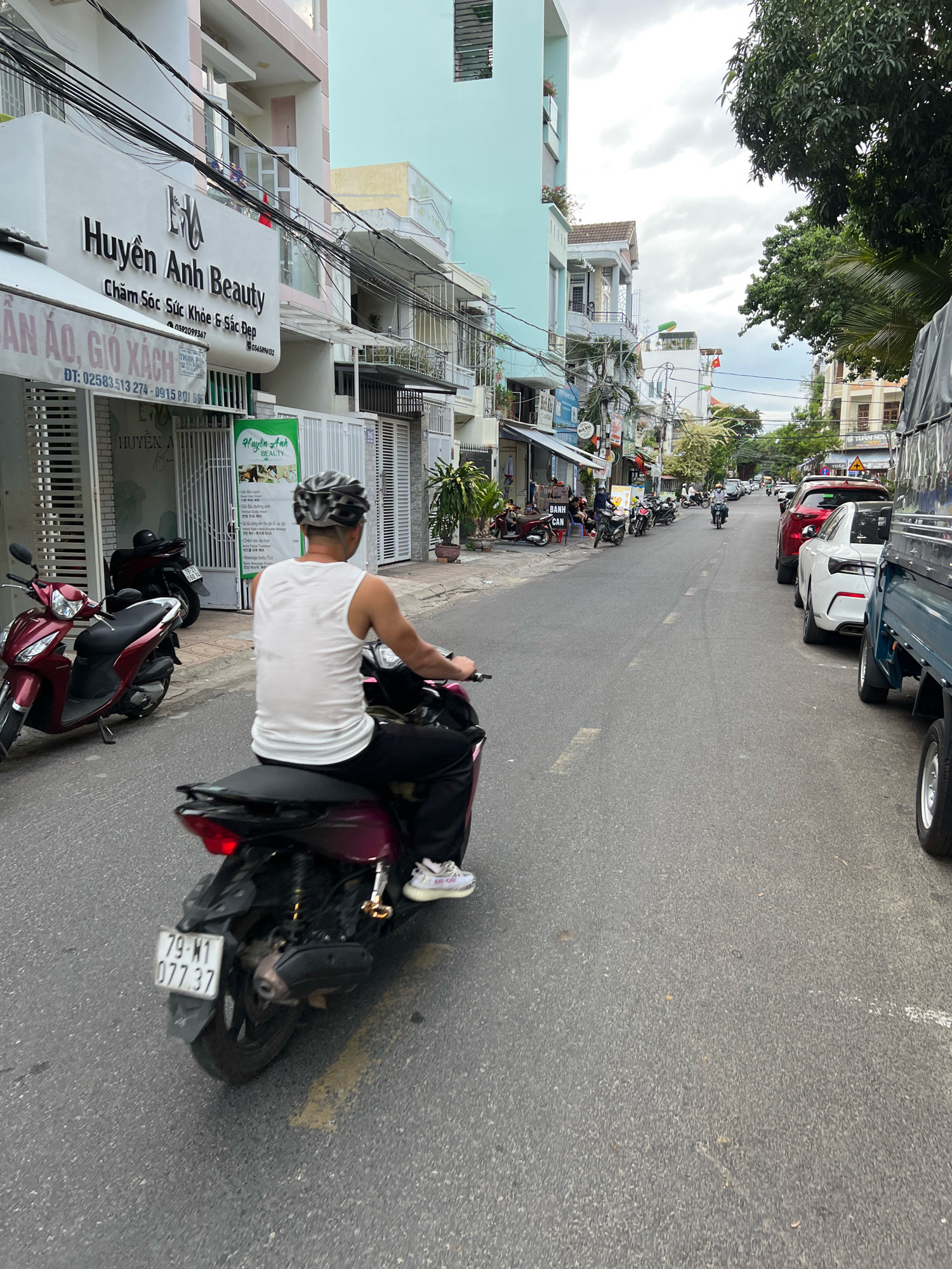 A man rides a scooter down a city street in Nha Trang, Vietnam. The man is wearing a white tank top, black pants, a helmet, and sneakers. He is seen from behind, with the scooter's license plate visible. The street is lined with buildings, including shops and apartments, and several other vehicles, including cars and scooters, are parked or driving on the road. The sky is cloudy. The buildings on the left side of the street have signs in Vietnamese. One sign reads "Huyen Anh Beauty" and lists phone numbers. Another sign reads "An Ao, Gio Xach" which translates to "Clothes, Handbags." The scene depicts a typical urban environment in Vietnam.