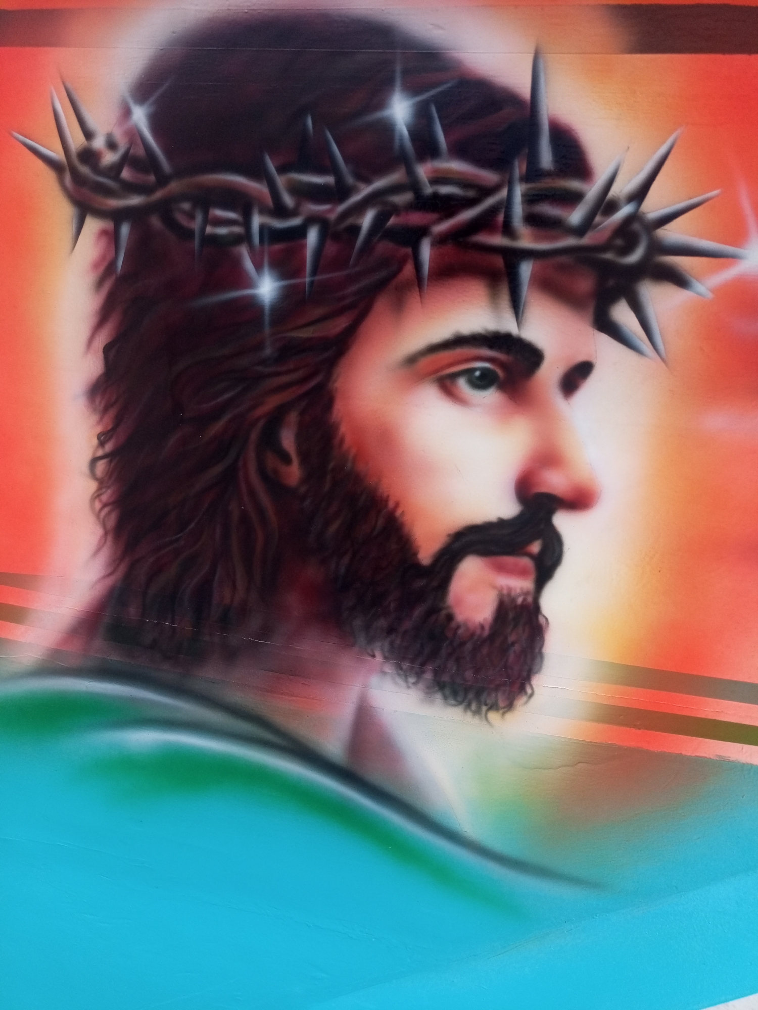 The image depicts a spray-painted mural of Jesus Christ wearing a crown of thorns. The background is a gradient of orange and red, with horizontal stripes of color running through it. The lower portion of the image is painted in shades of blue and green. The mural is likely located in the city of Juan Griego, Venezuela.