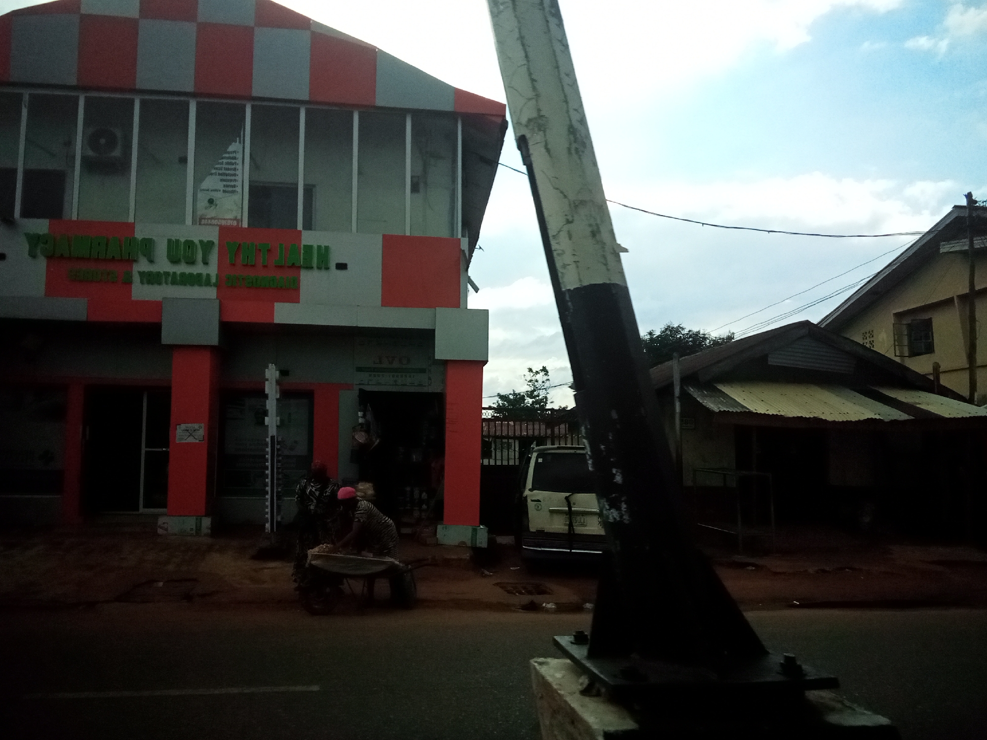 In the city of Nibo, Nigeria, this image captures a street scene dominated by a two-story building with a checkered red and gray facade. The building features large windows and prominently displays signage in green lettering that reads "HEALTHY YOU PHARMACY." Below this, smaller green text indicates "CONSULTING & LABORATORY SERVICES." To the right of the pharmacy, a corrugated metal-roofed structure with an open front and some display shelves is visible. A white van is parked in front of this structure. In the foreground, near the pharmacy entrance, two individuals are present; one is pushing a wheelbarrow filled with goods, while the other appears to be attending to its contents. A utility pole, partially obscuring the view, stands in the right foreground. The sky above is partly cloudy, with the sun casting soft light on the scene, indicating it's likely daytime. The ground in front of the buildings appears to be a dirt road or pathway.