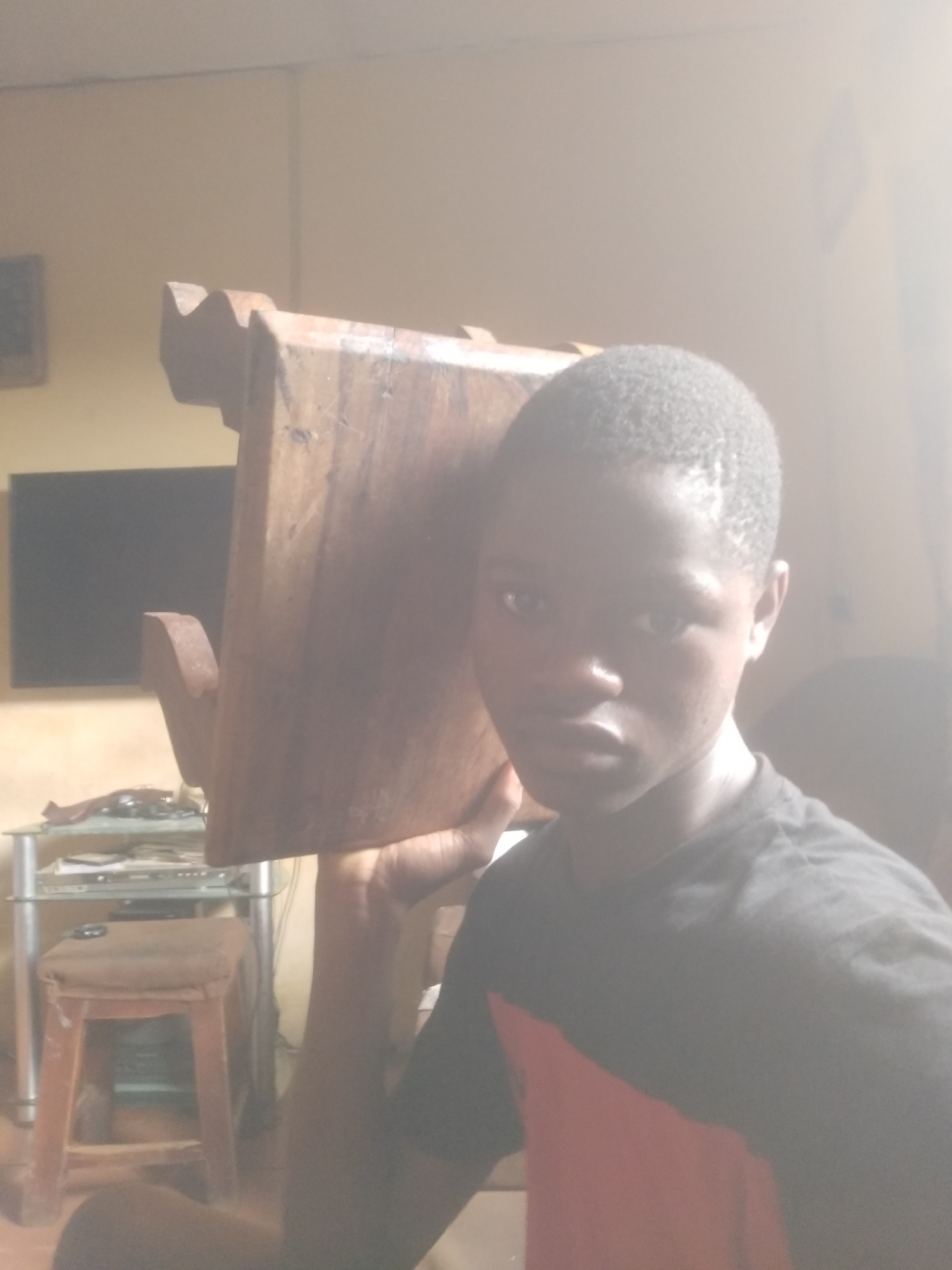 A person holds a wooden object, possibly furniture, at shoulder level. The person has short, dark hair and is wearing a dark t-shirt with a red patch. Background elements include a TV, a glass-topped table with items on it, and a wooden stool. The location appears to be indoors, with light streaming in from the side. The city and specific location are unknown.