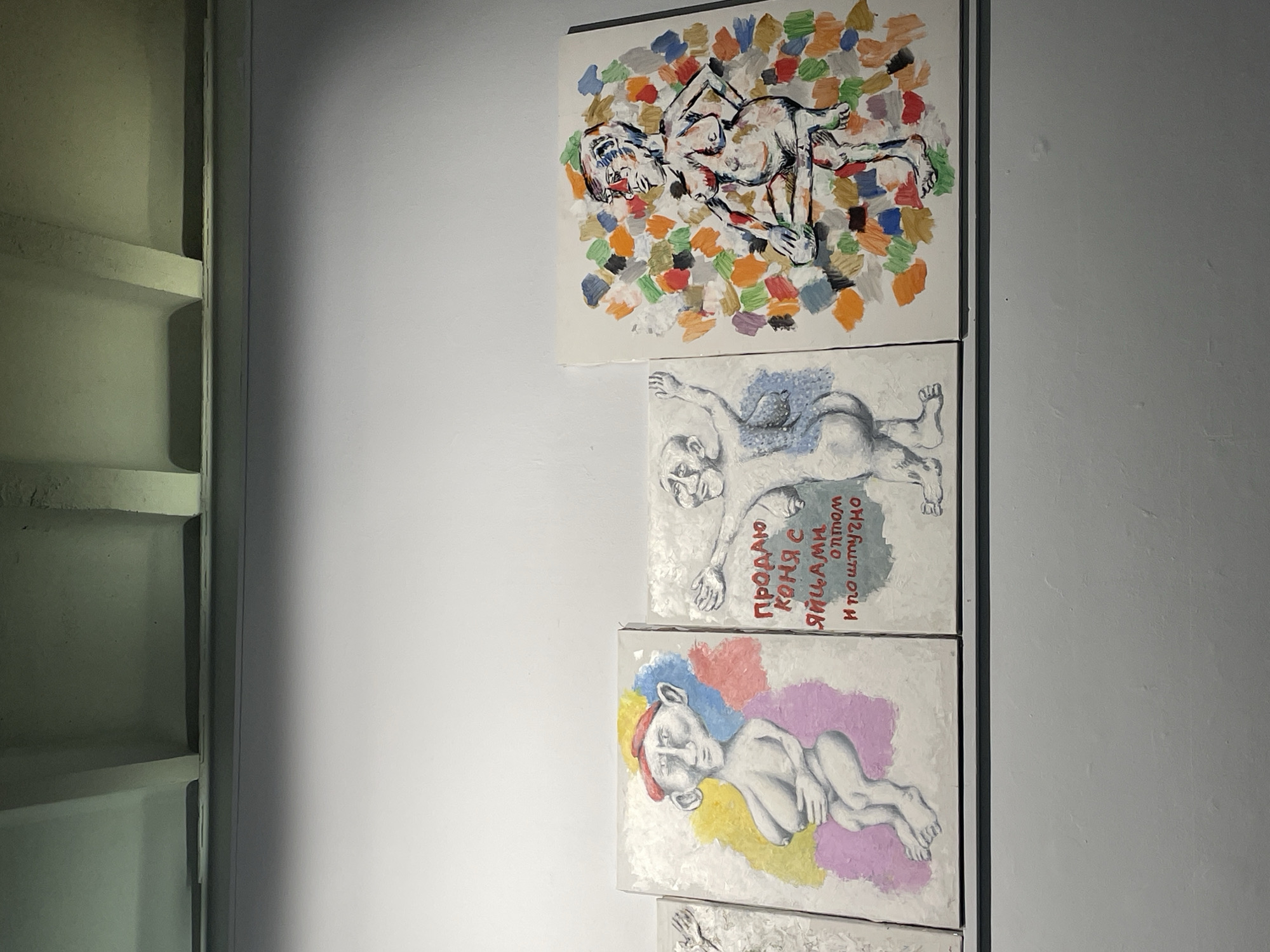 This is a vertical shot of several paintings displayed on a wall. The paintings are arranged in a column. The leftmost part of the image shows a section of what appears to be a wall with built-in shelving. The right side of the image shows a light gray wall where the artworks are mounted.
The top painting depicts a stylized, abstract human figure in a reclining position. The figure is outlined in black and filled with a mix of white and pink, with some dark lines suggesting musculature. The background is a collage of multicolored, roughly painted squares in shades of green, orange, blue, red, purple, and brown.
Below the first painting is a second artwork, a monochromatic painting of two figures. One figure is larger and lying down, with its head turned to the side and its arms extended. A smaller figure is partially visible behind the larger one, also in a reclining pose. The background is a textured white with some blueish-gray elements. There is visible text in Cyrillic script on this painting, which reads: "ПРОДАЮ КОНЯ С ЯЙЦАМИ оптом и поштучно" (translates to "SELLING A HORSE WITH BALLS wholesale and retail").
The third painting features a figure that appears to be a child or a young person, lying on their side. The figure is rendered in shades of gray and white. The background is a vibrant splash of colors including yellow, red, blue, and purple.
The bottom of the image shows a sliver of a fourth painting, similar in style to the second one, depicting a person's head and shoulders.
The lighting in the scene is somewhat dim, suggesting an indoor setting like an art gallery or studio. The overall mood is artistic and slightly unsettling due to the subject matter and the abstract style of some of the paintings.