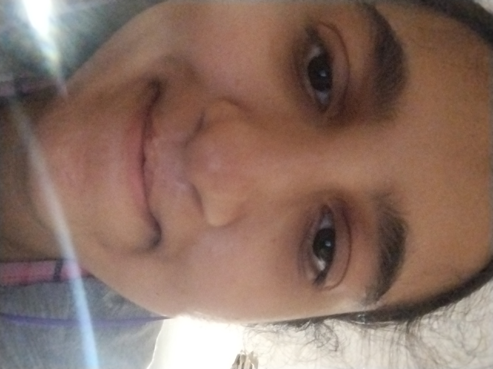 The image is a close-up, over-the-shoulder selfie of a young female with dark hair and brown eyes. She is wearing a grey shirt and a purple lanyard. The background is blurry and appears to be an indoor setting with bright lighting. There are no distinct landmarks or environmental features visible to determine the location in Plachkovtsi, Bulgaria. The subject is looking directly at the camera with a neutral expression.