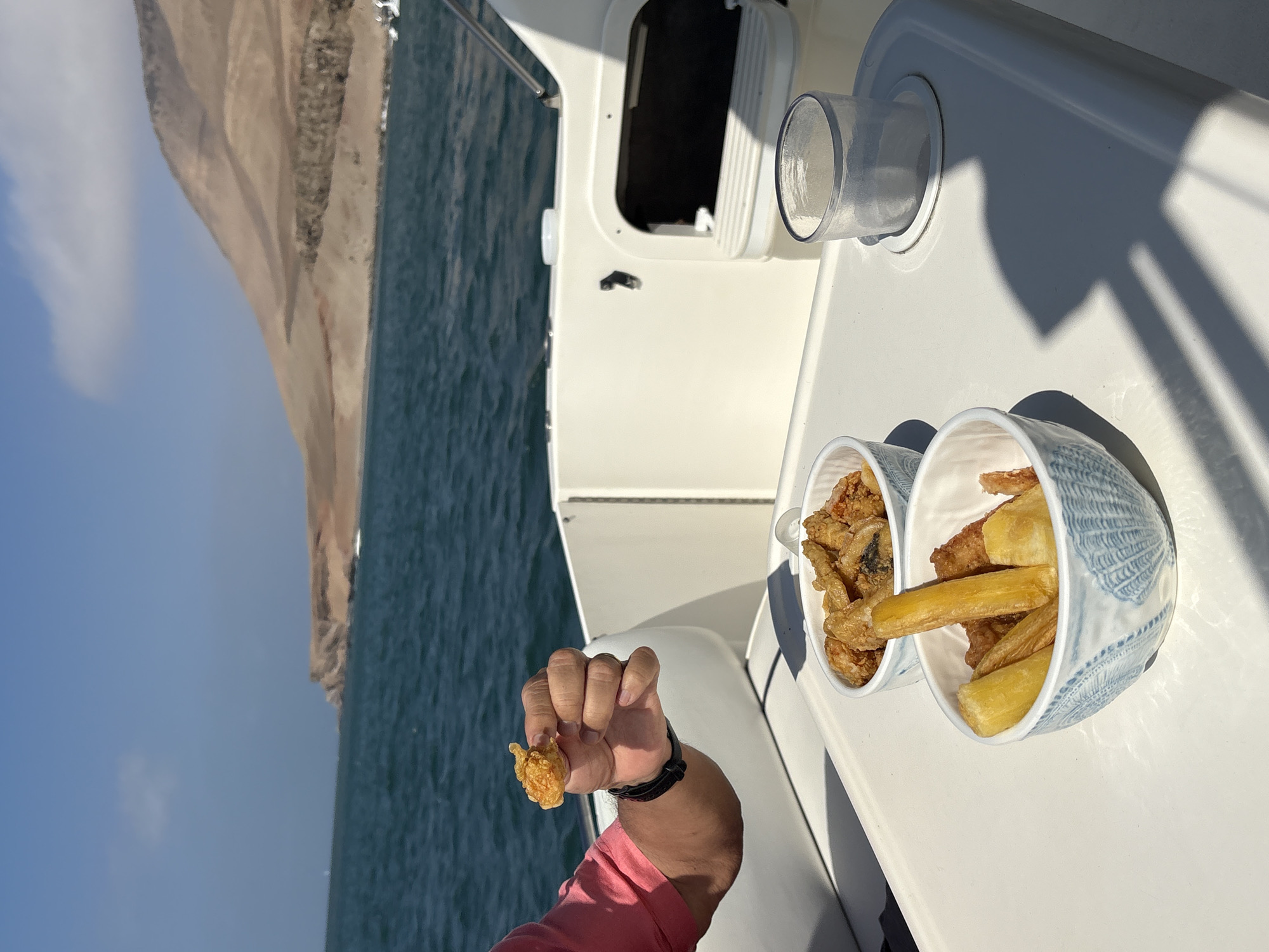 On the deck of a boat, an arm wearing a black wristband holds a piece of fried food. Beside it, there are two bowls: one containing fried plantains, and the other holding a mix of fried seafood. The setting appears to be near a coast, possibly the city of Callao, Peru, as suggested by the coastal landscape. The water stretches out towards a tall, beige rock formation on the left. The boat is white and shows signs of outdoor use. The day is sunny, casting clear shadows.