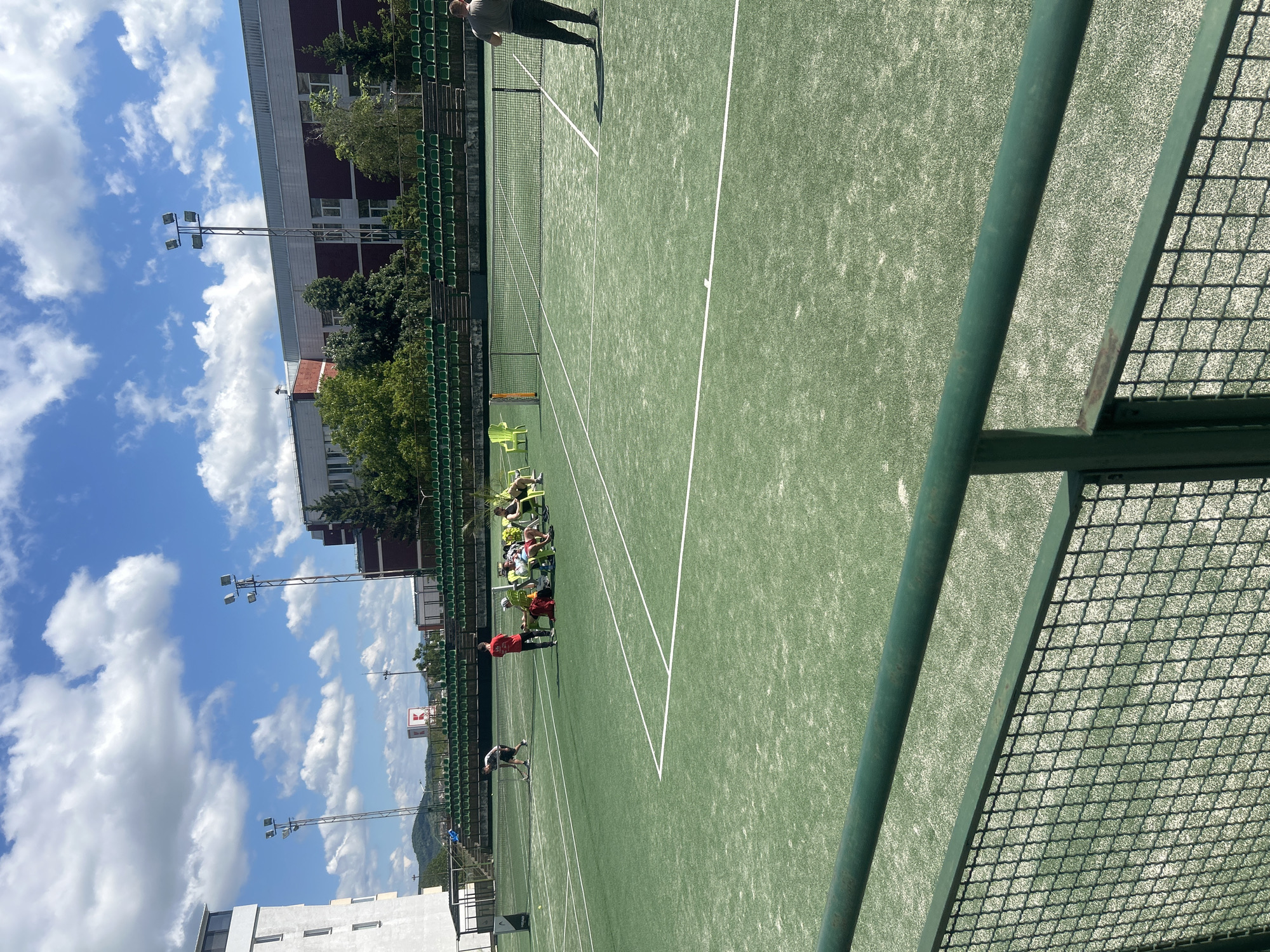 The image shows a tennis court in Veliko Tarnovo, Bulgaria. The court has a green surface with white lines. A tennis net divides the court in half. There are several people on the court. Some of them are sitting on chairs near the back of the court, and others are standing and appear to be playing tennis. The background includes a building with green stadium seating, trees, lamp posts, and a blue sky with scattered clouds. A green metal fence is visible in the foreground.