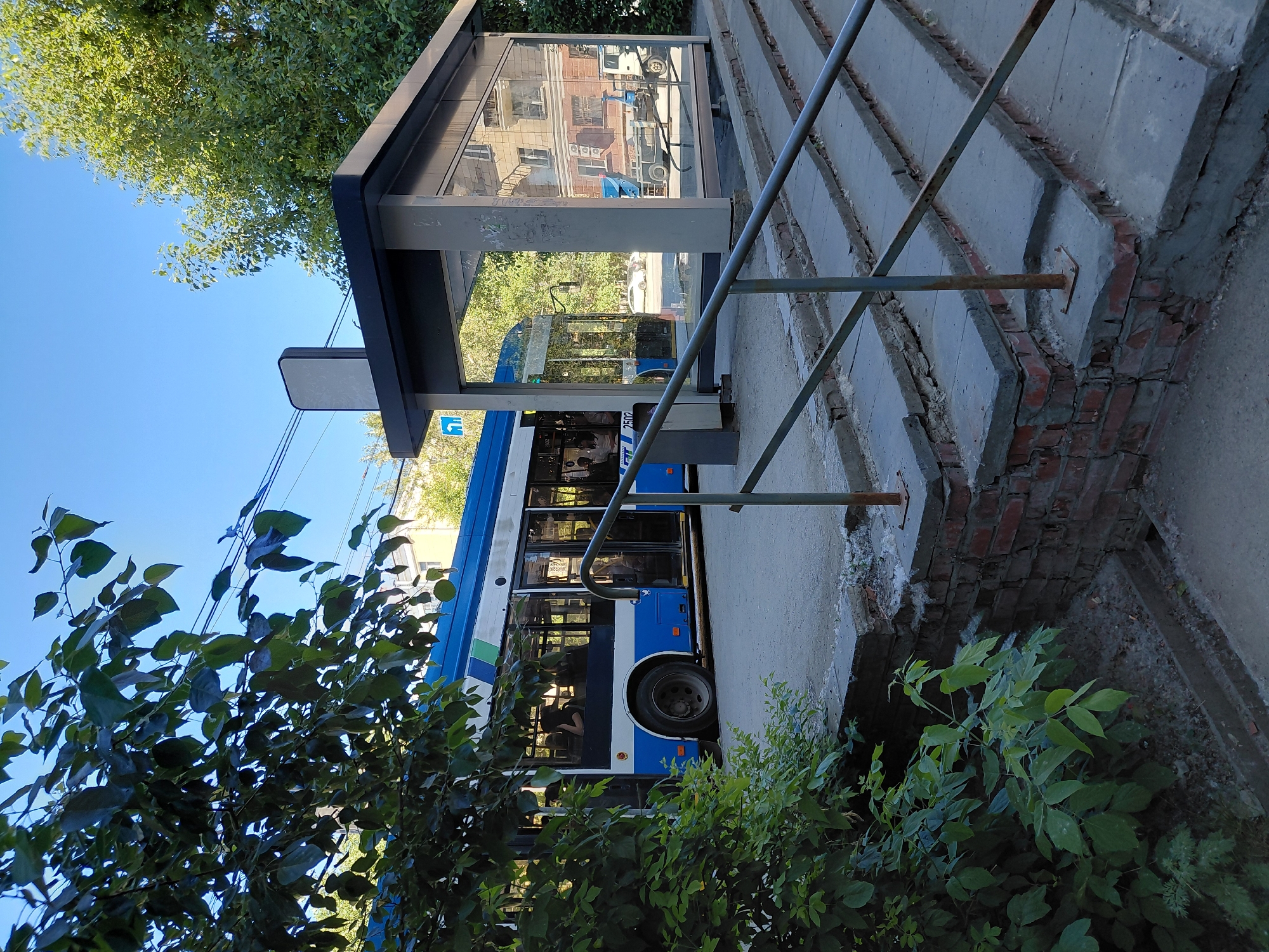 An outdoor, eye-level shot captures a city street scene, dominated by a bright blue and white bus stopped at a bus stop. The bus, with its doors open, has passengers visible inside. The bus stop itself is a modern structure with glass walls and a dark roof, reflecting the surrounding environment. In the foreground, a weathered brick wall and concrete steps lead up to the bus stop, with a metal handrail providing support. Lush green foliage from a tree partially obscures the left side of the image. The sky above is a clear, bright blue, suggesting a sunny day. The background reveals buildings and more trees, indicating a developed urban setting.