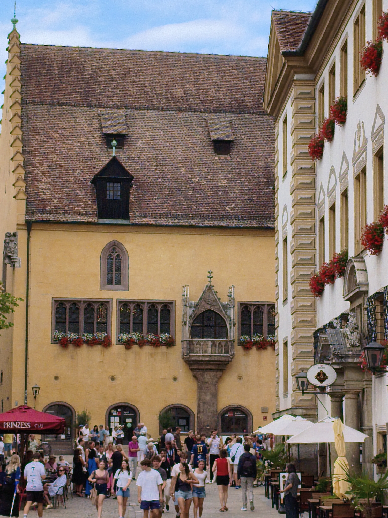The image depicts a bustling outdoor scene in the city of Regensburg, Germany, likely in the historic city center given the architectural style. A large, ornate yellow building with multiple windows and a tiled roof dominates the left side of the frame. To the right, a lighter-colored building with decorative elements and flower boxes is visible. A crowd of people is present, walking and gathered in small groups. Some individuals are seated at tables outside, suggesting a cafe or restaurant setting. The overall atmosphere appears lively, with people enjoying a day outdoors. The sky is partly cloudy.
