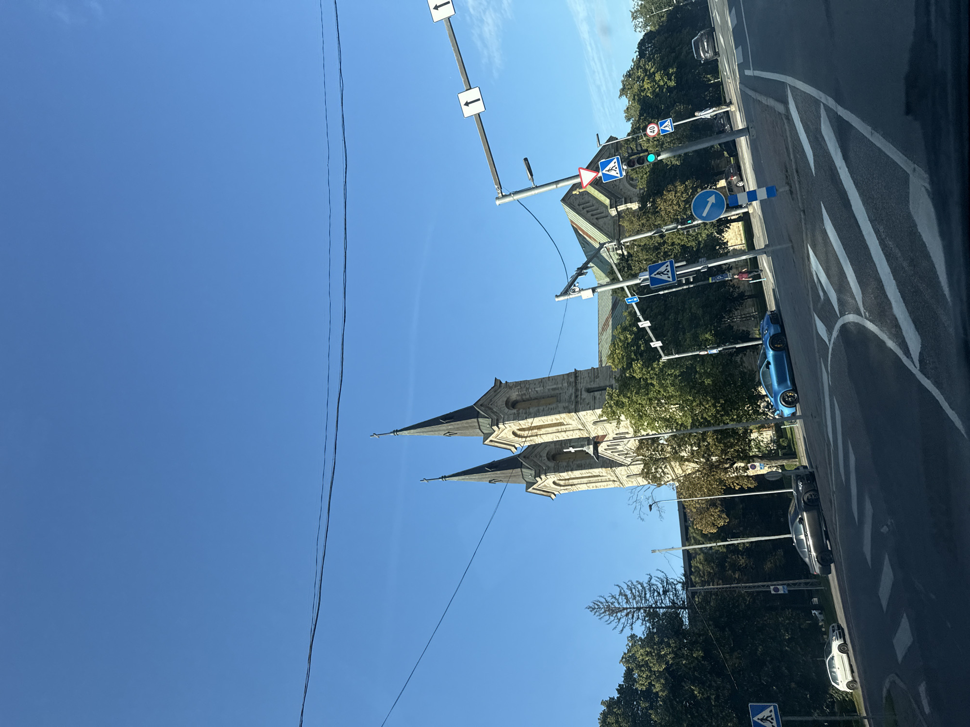 The image depicts a view of the St. Peter and Paul Cathedral in Tallinn, Estonia, from a street level perspective. The grand stone cathedral, characterized by its twin spires topped with crosses, stands prominently against a clear blue sky. Dense green trees flank the cathedral, suggesting a park or a well-landscaped area surrounding the religious edifice.
Several traffic signs are visible on traffic light poles, indicating street regulations and directions. These include pedestrian crossing signs, a yield sign, and a speed limit sign displaying "40". Traffic lights are also present, showing green signals. A blue sports car is parked near the roadside, and other vehicles, including a white car and a dark sedan, are observed on the road. The foreground shows a street with clearly marked pedestrian crossings, indicating an intersection or a busy road. The overall scene captures a moment in the city life of Tallinn, with a blend of historical architecture and modern urban infrastructure.
