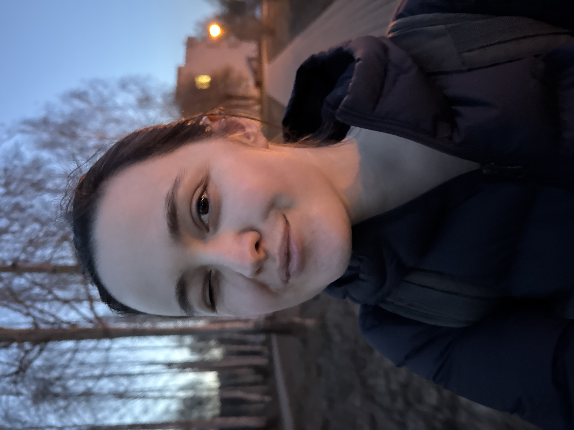 A young woman is taking a selfie outdoors at dusk. She is wearing a dark blue puffer jacket. The background includes bare trees, a dark wooden fence, a lit building, and a street lamp. The specific location in Izhevsk, Russia, cannot be determined based on the provided visual information.