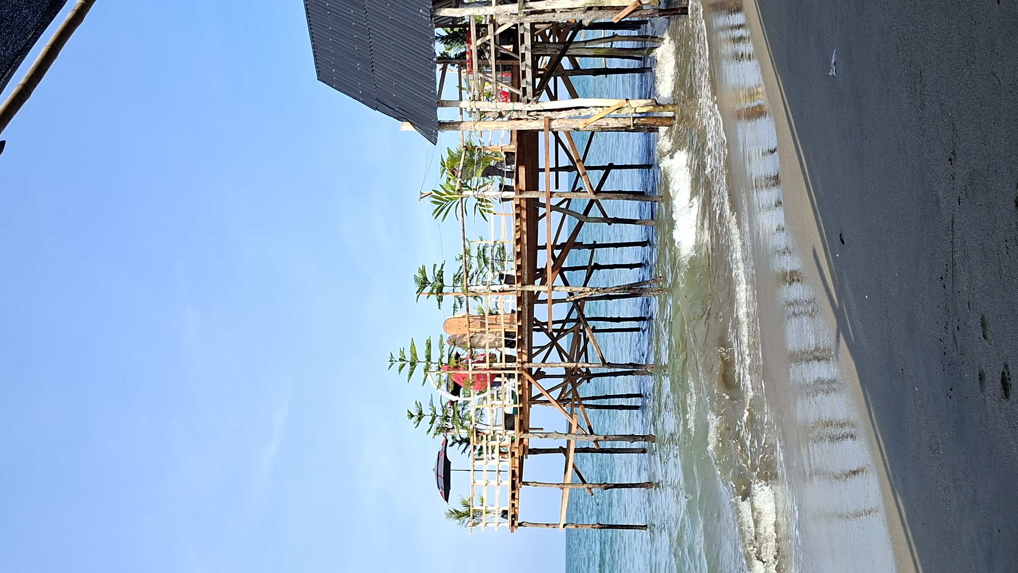 A wooden pier extends over the turquoise water at a beach in Lhokseumawe, Indonesia. The pier is supported by numerous wooden stilts driven into the seabed. On the pier, several potted plants, including pine trees and palms, are arranged along a wooden railing. Two individuals are visible on the pier; one, wearing a red shirt, stands near a black umbrella, while the other, in a light-colored shirt, stands further down the pier. A surfboard leans against the railing. To the right of the pier, a small structure with a dark, sloped roof is partially visible. Gentle waves are breaking on the sandy shore in the foreground, and the reflection of the pier's stilts is visible on the wet sand. The sky above is clear and a bright blue.