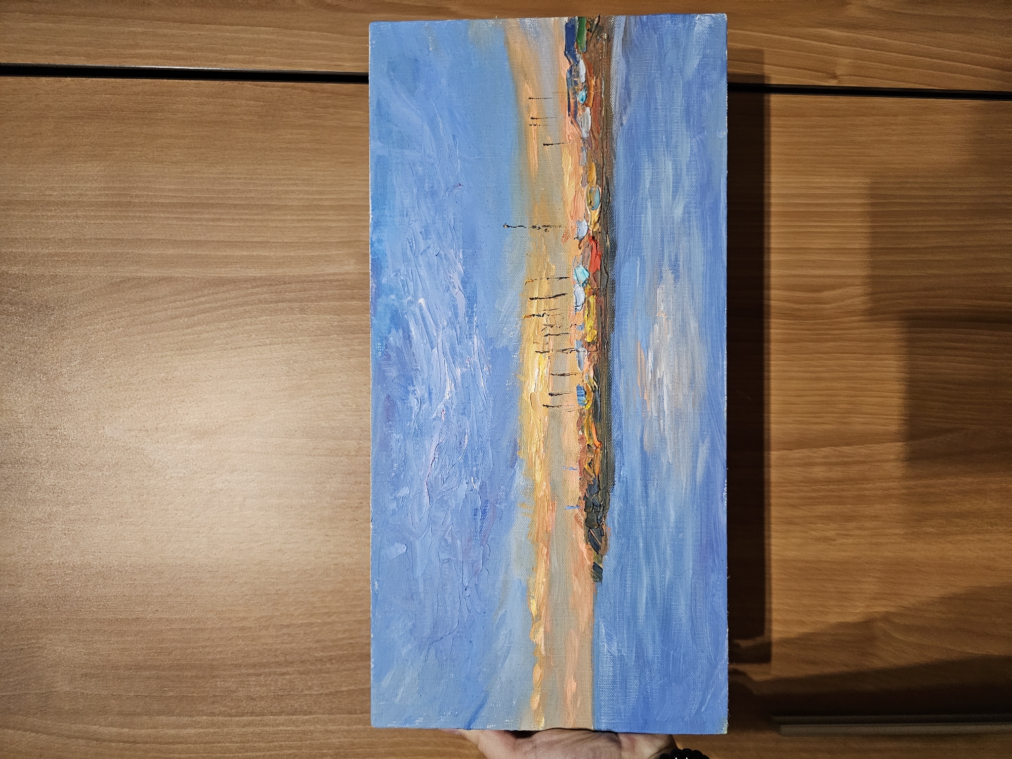 The image shows a rectangular oil painting held by a person against a wood-grain background, likely a wall or furniture. The painting depicts a coastal scene, divided vertically into three primary sections. The left and right sides are dominated by varying shades of blue, representing the sky and the sea respectively, with visible brush strokes creating texture. The central section features a strip of land with colors ranging from yellow and orange to brown, suggesting a beach or coastline at sunset or sunrise. There are also hints of structures or objects along the coastline, including what appears to be colorful umbrellas. The painting has a textured, impasto effect, with thick layers of paint applied.
The location in Plovdiv, Bulgaria is not represented in the painting. The painting is depicting a coastal scene.