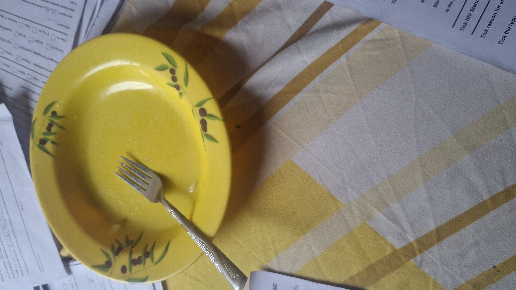 A close-up, overhead shot shows a yellow, round plate with a fork resting inside it. The plate is decorated with a simple pattern of olive branches. The plate is positioned on a checkered tablecloth with diagonal yellow and white stripes. Several sheets of paper are visible in the background, some with handwritten text and checkboxes. The lighting appears to be natural, casting soft shadows. The overall scene suggests a casual setting, possibly a dining area.