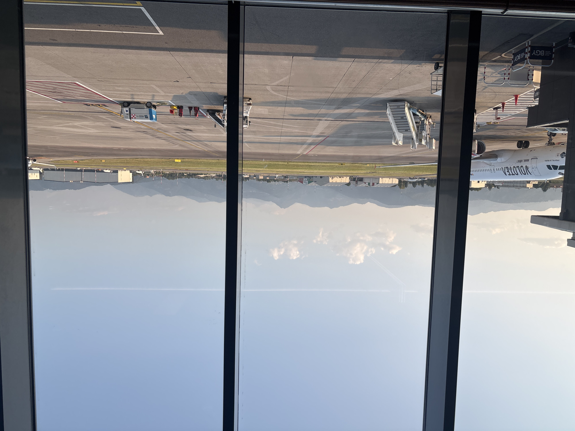 An outdoor, daytime, wide shot depicts an airport tarmac with a clear sky and distant mountains. In the foreground, through a large window with dark vertical frames, an airplane with "VOLOTEA" branding is parked. A mobile boarding staircase is positioned next to the aircraft, and baggage carts are visible. Further down the tarmac, other aircraft and airport buildings, including hangars, are visible. The scene is characterized by the bright sunlight casting long shadows across the concrete. The location is Orio al Serio, Italy, specifically the Bergamo Airport (BGY). There are no people clearly visible in the image.