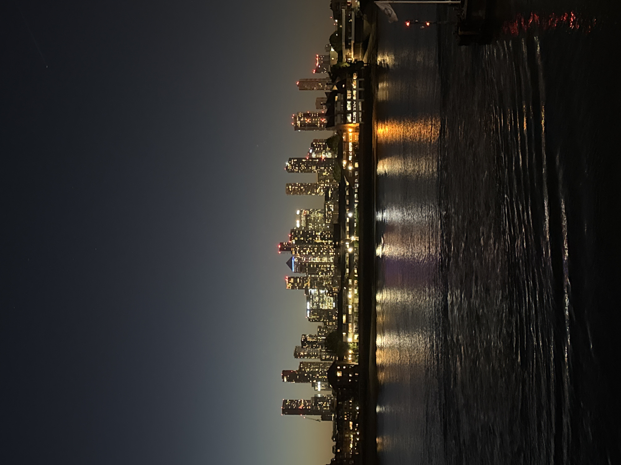 This is a night-time landscape image depicting a modern cityscape in London, United Kingdom, viewed across a tranquil body of water, likely the River Thames. The horizon is dominated by a dense row of numerous high-rise buildings and skyscrapers, brightly illuminated by the many lights from their windows. These lights create a warm, vibrant glow, with several structures also featuring distinct red lights near their summits.
The artificial illumination from the city skyline casts striking reflections onto the dark, rippled surface of the water. Streaks of yellow, orange, and white light are stretched and distorted across the water, mirroring the urban luminescence above. The water appears calm, suggesting fair weather conditions.
The sky above is a deep, dark blue, gradually lightening towards the horizon, typical of a late evening or early morning scene. A few faint, small points of light can be seen in the upper sky. On the far right, a section of a pier or dock is visible, marked by additional red lights. A flag on a flagpole is partially visible in the upper right corner.
No specific activities or individuals are captured in the frame, presenting a serene yet dynamic urban vista. There is no discernible text within the image. The photograph conveys the captivating beauty and nocturnal energy of a major metropolitan area.