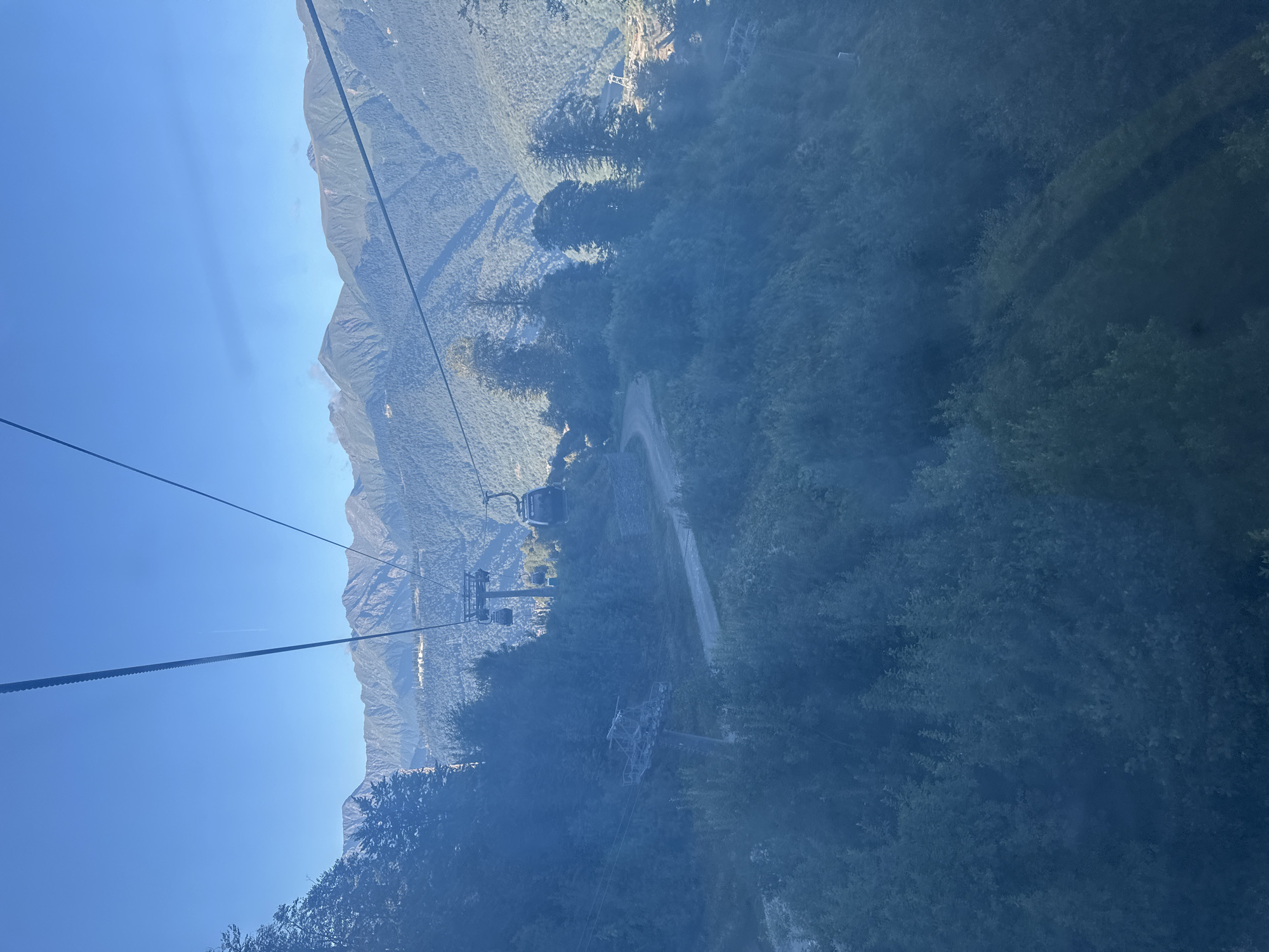 The image captures a view from a cable car ascending a mountain in Estosadok, Russia. The cable car itself is partially visible in the foreground, as are the thick cables supporting it. Below, the mountain slopes are covered in dense green foliage, with a winding dirt road visible through the trees. In the distance, several other cable car cabins can be seen, along with the imposing peaks of the Caucasus Mountains under a clear blue sky. The overall scene depicts a serene mountain landscape, likely a popular tourist destination during warmer months.