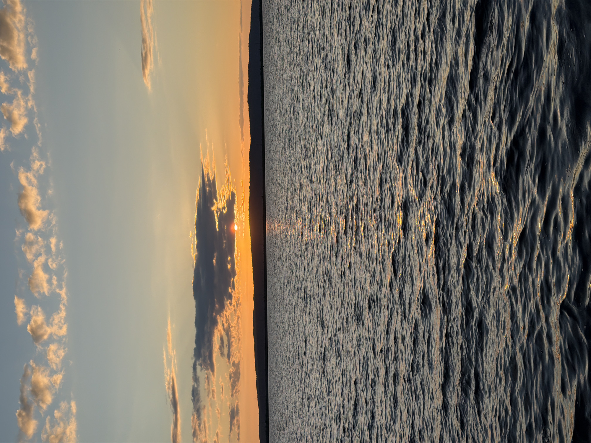 The image depicts a tranquil scene of a sunset over a body of water, likely the Kama River, in Chaikovsky, Russia. The sun, partially obscured by clouds, casts a warm orange glow across the sky and reflects on the rippling surface of the water. The horizon is marked by a dark silhouette of hills and trees, with faint power lines visible against the sky. The sky transitions from a pale blue at the top to a vibrant orange and yellow near the horizon, with scattered clouds catching the last rays of sunlight. The water is dark and choppy, creating a textured foreground. No people or specific objects are identifiable in the scene.