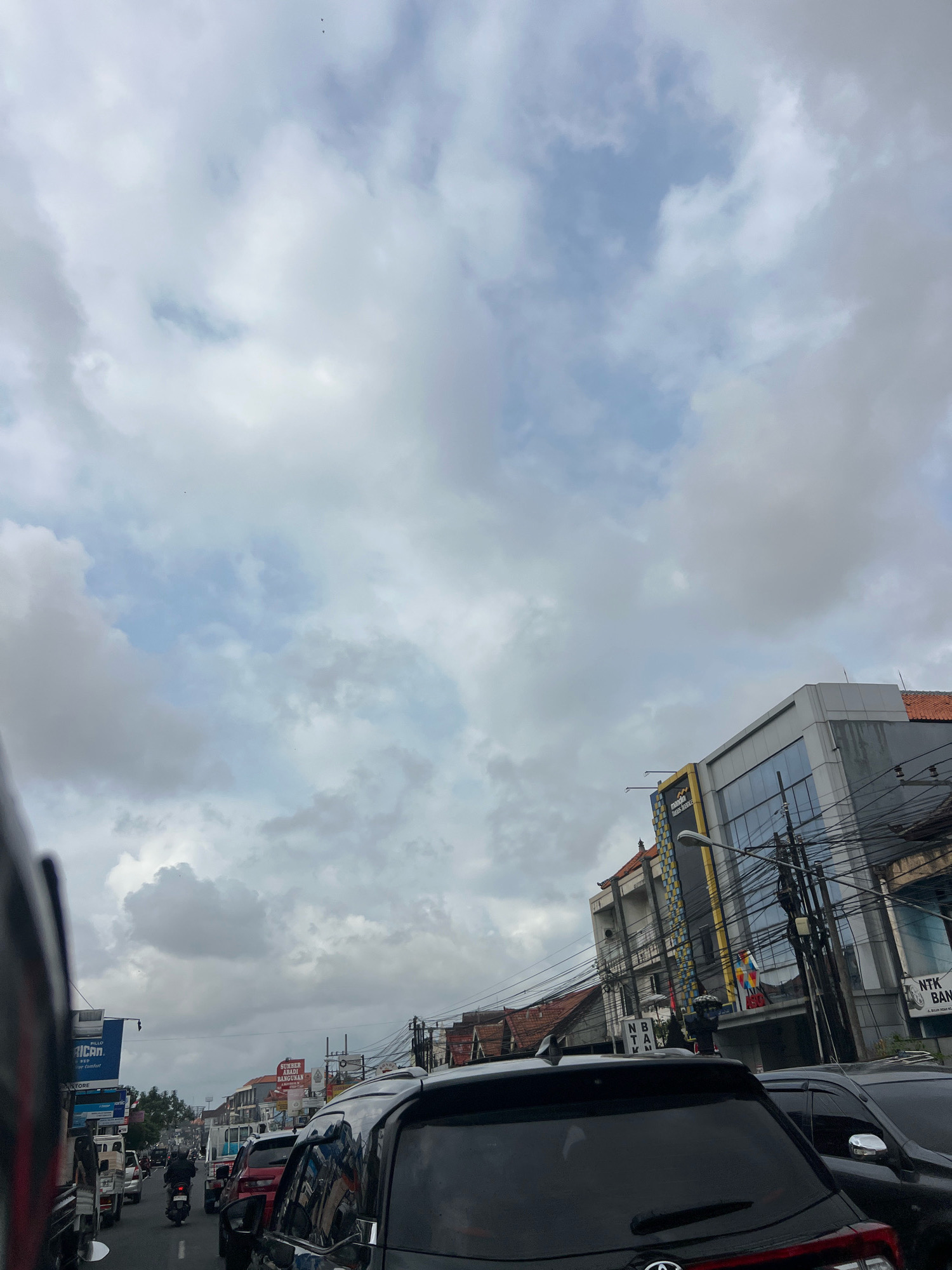 The image captures a street scene in Denpasar, Indonesia, with a focus on a traffic jam under a cloudy sky. In the foreground, the rear of a black SUV is prominently visible, reflecting the surrounding environment. To the right, another vehicle, also dark in color, is parked. Further down the road, a line of cars, including a white truck and a red car, are stationary. A motorcycle with a rider is also part of the congested traffic.
The urban environment is characterized by buildings lining the street. One building on the right features a modern glass facade with signs, including "Mandiri Bank" and "NTB Bank." Another building to the left displays a blue billboard for "Pillars." Telephone wires are strung across the scene, adding to the urban feel. The sky is filled with thick, gray clouds, suggesting overcast weather. The overall impression is one of a busy city street during a traffic slowdown under a moody sky.
