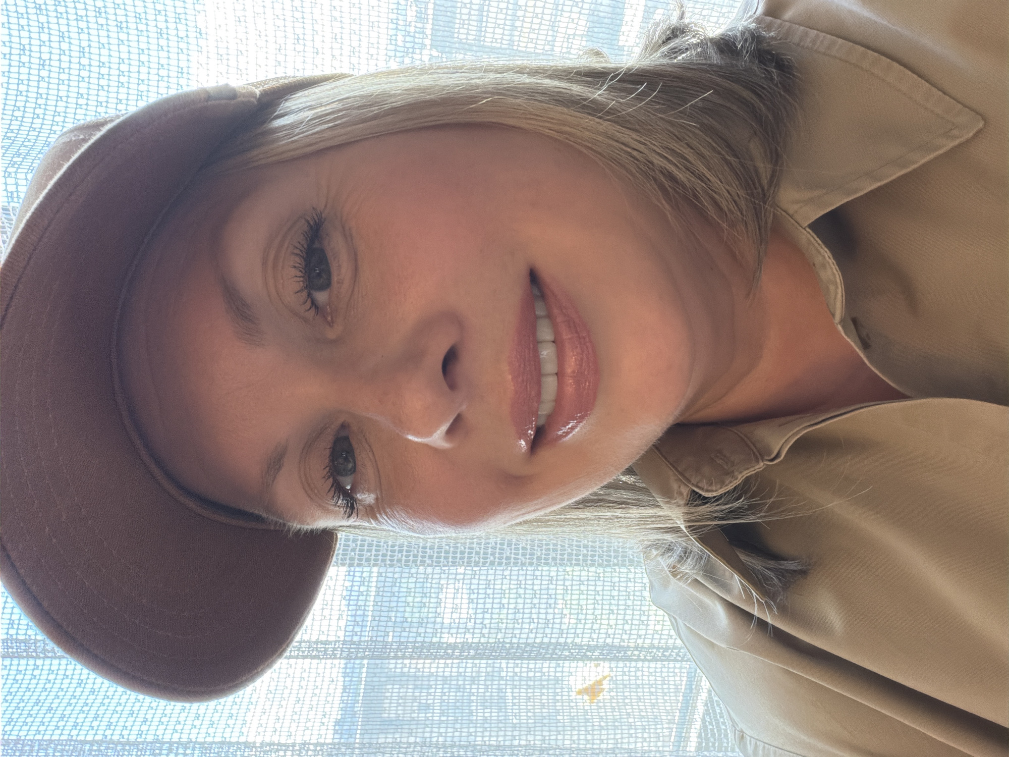 A woman with blonde hair and wearing a brown cap and a tan collared shirt is looking directly at the camera. She has on makeup, including eyeliner, mascara, and lip gloss. The background appears to be a window with sheer white curtains, and some blurry outdoor scenery is visible through them.