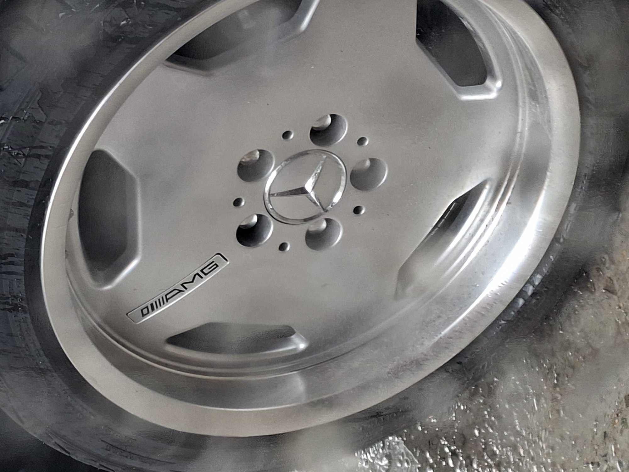 The image shows a close-up view of a silver Mercedes-Benz AMG wheel with the AMG logo visible. The wheel is partially covered by a wet tire. Water and spray are visible around the lower portion of the wheel and tire, suggesting it is being washed or has just been washed. The background is blurred but appears to be an outdoor, possibly urban, environment with wet ground. The scene is consistent with a car wash or a street in Barnaul, Russia, during wet weather conditions. There are no people or direct interactions visible in the frame.