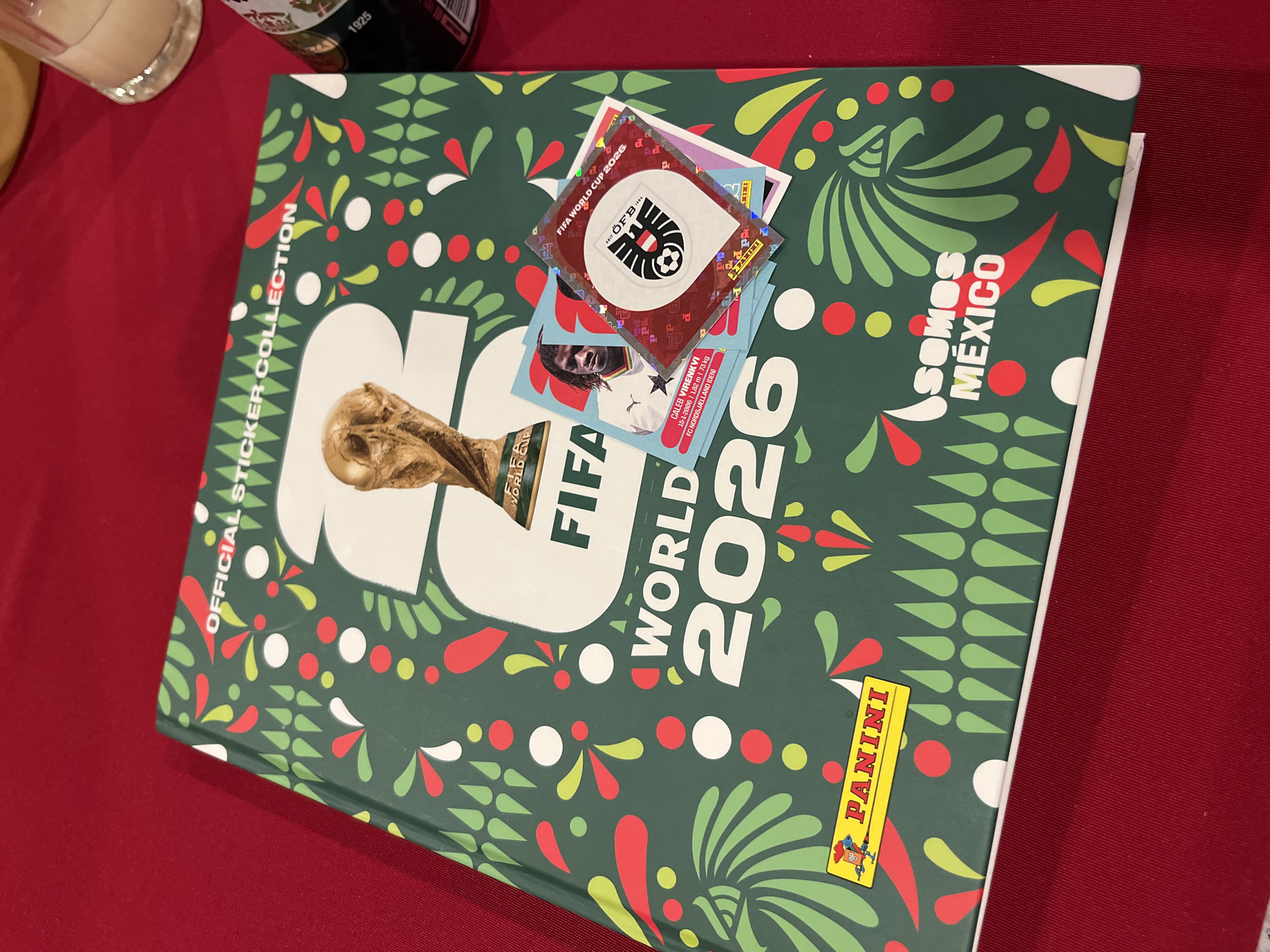 The image shows a FIFA World Cup 2026 official sticker collection album lying on a red surface, possibly a tablecloth. The album has a green cover with decorative floral and geometric patterns in red, white, and yellow. The words "Somos México" are printed on the upper right corner of the album cover. Several FIFA World Cup stickers are stacked on top of the album. The stickers include one with the Österreichischer Fußball-Bund (ÖFB) logo and another with the player Caleb Virenkvi. Part of a beer bottle and a glass are visible in the upper left corner. The location and city information are not evident in the image.