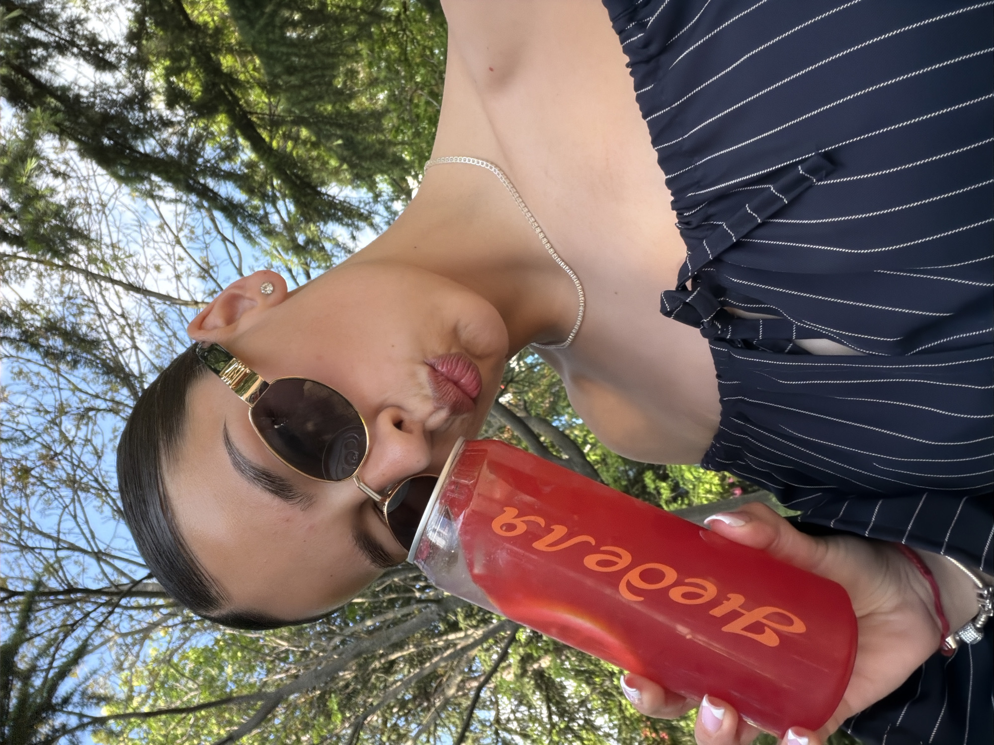 A young woman with sunglasses and a necklace is drinking from a red plastic can. She is wearing a navy blue and white striped top. The background shows trees and sky. It is not possible to determine if the location is Kazanlak, Bulgaria without additional information.