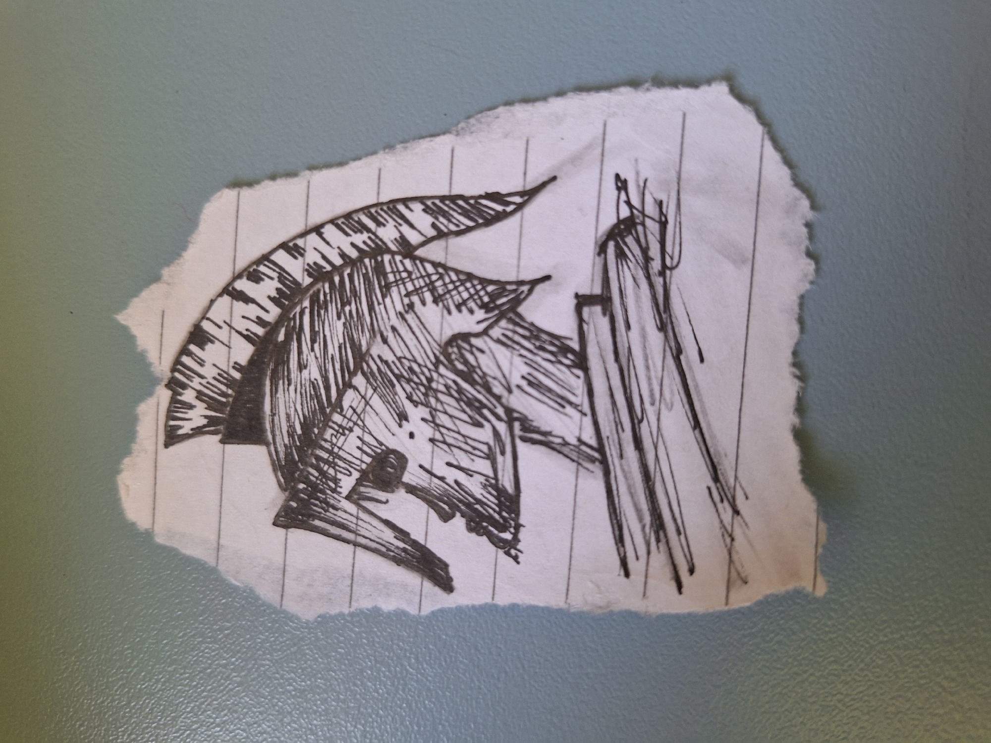 A torn piece of lined notebook paper with a black ink sketch of a Spartan warrior's helmet and upper body is centered on a pale blue, textured surface. The sketch, which appears to be from a profile view, shows the characteristic plumed helmet, a portion of a face with defined cheekbones and an eye socket, and the beginning of a neck and torso. The ink lines are varied in thickness and density, creating shading and texture. The paper is irregularly torn along its edges. The blue surface has a subtle, pebbled texture. There are no people or animals present in the image, and no specific Varna landmarks are visible.