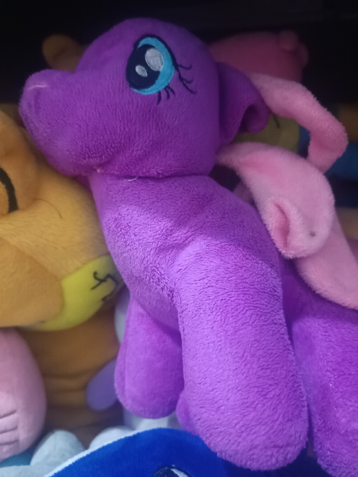 The scene, situated within the city of Gotha, Germany, depicts a close-up view of multiple plush toys. The central subject is a medium purple plush toy, characterized by large, embroidered eyes featuring black pupils, white highlights, light blue irises, and three delicate black eyelashes. It possesses small, rounded purple ears and a soft snout. Two light pink plush wings are partially visible on its back, one curving upwards and the other downwards. The toy's body and a single visible leg are also purple. The texture throughout is a short-pile, fuzzy plush fabric. To the left, a portion of a yellowish-orange plush toy is visible, displaying a light yellow area with faint dark embroidered markings. In the background, indistinct pink plush elements are discernible. At the bottom of the frame, a segment of a royal blue plush toy is present, featuring a visible black embroidered eye with a white highlight, mirroring the style of the central purple toy's eyes. The plush toys are densely arranged, suggesting a confined display or storage environment.
