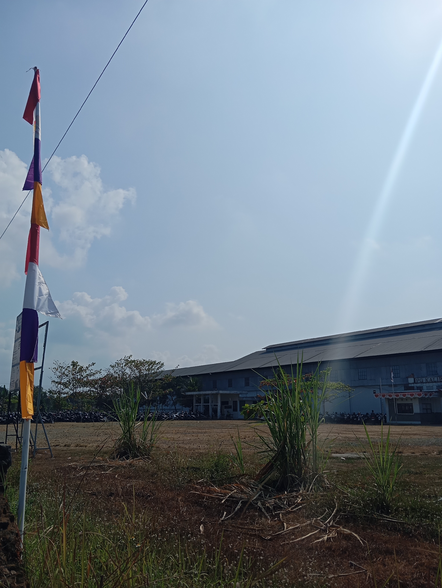 The image shows a clear sky with a bright sun, casting a lens flare on the right side. In the foreground, a tall pole flies several colorful flags, including red, white, purple, and yellow. The flags are triangular and appear to be made of fabric. To the right of the flags, there is dry, brown grass and some tall green plants. In the background, a long, grey building with a dark roof stretches across the frame. The building has several windows and a white facade on the lower level. In front of the building, a large number of motorcycles are parked in rows. The scene appears to be an open area outside the building, possibly a parking lot or a courtyard. The building has signage that partially reads "PG KALIB." This strongly suggests the location is PG Kalibendo, a sugar factory in Banyumas Regency, Central Java, Indonesia.
