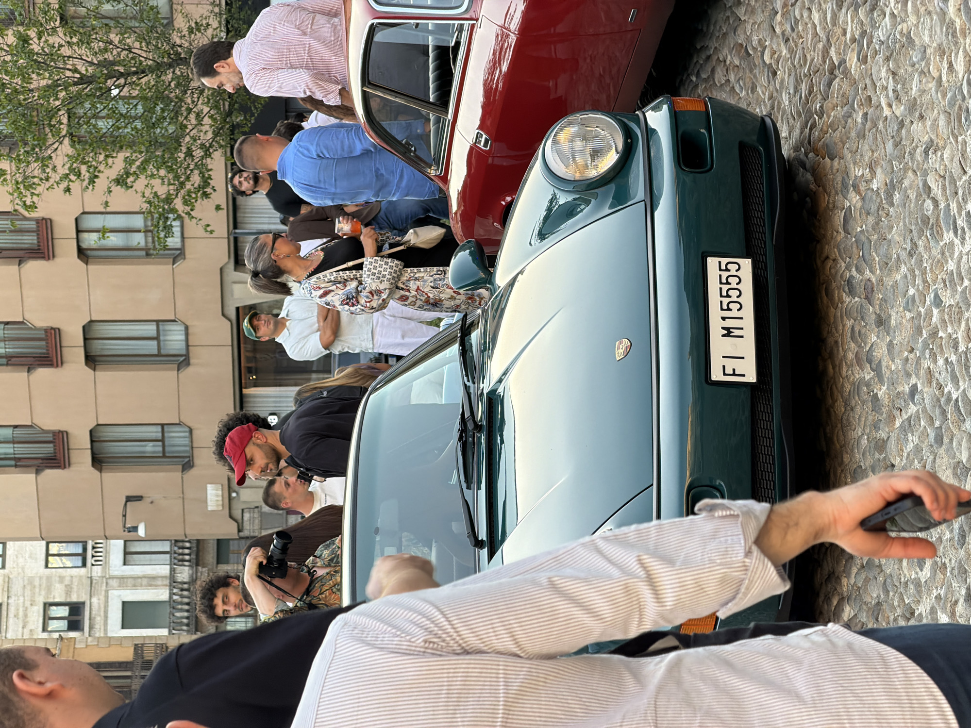 In this image, a group of people are gathered around two classic cars. The car in the foreground is a dark green Porsche, prominently displaying its license plate which reads "FI M15555". Beside it, partially visible, is a red car. The scene appears to be taking place outdoors, with a cobblestone street surface visible. The people are dressed in casual attire, and some are engaged in conversation or observing the cars. One person is holding a camera, suggesting the event might be a car show, gathering, or a photo opportunity. The lighting suggests it could be late afternoon or early evening. The backdrop includes buildings with windows, typical of an urban environment. The overall atmosphere seems relaxed and social, with individuals showing interest in the vintage vehicles.