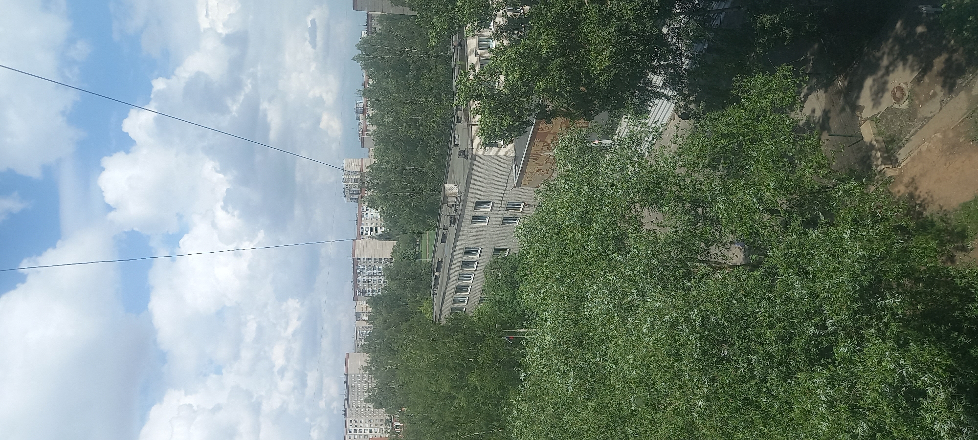 This is a high-angle, full shot of a residential area in Izhevsk, Russia, on a partly cloudy day. The foreground is dominated by lush green trees with dense foliage, partially obscuring the view of buildings behind them. A closer building, appearing to be an apartment complex, is visible in the mid-ground with a grey facade, rows of windows, and a flat roof. In the background, multiple high-rise apartment buildings stretch across the horizon, characterized by their neutral color palettes and a typical Soviet-era architectural style. The sky above is a mix of blue sky and white, fluffy clouds. Two thin, dark lines, likely wires or cables, diagonally traverse the sky from the top left to the bottom right of the frame. The scene depicts an urban environment with a significant presence of greenery, suggesting a well-treed neighborhood. There are no discernible people or active events captured in the image.