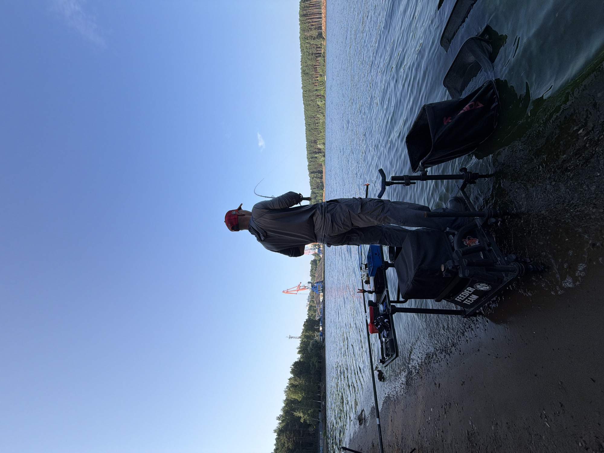 A man stands on the shore of the Kama River in Chaikovsky, Russia, fishing. He is wearing a red baseball cap, a gray hooded sweatshirt, and gray cargo pants. He holds a fishing rod, casting it into the water. In front of him is a fishing station with multiple rod holders, bait containers, and a seat. A black fishing bag is partially submerged in the river next to the station. The background features a dense forest on the opposite bank of the river and industrial cranes and structures in the distance, suggesting a port or industrial area. The sky is clear and blue.