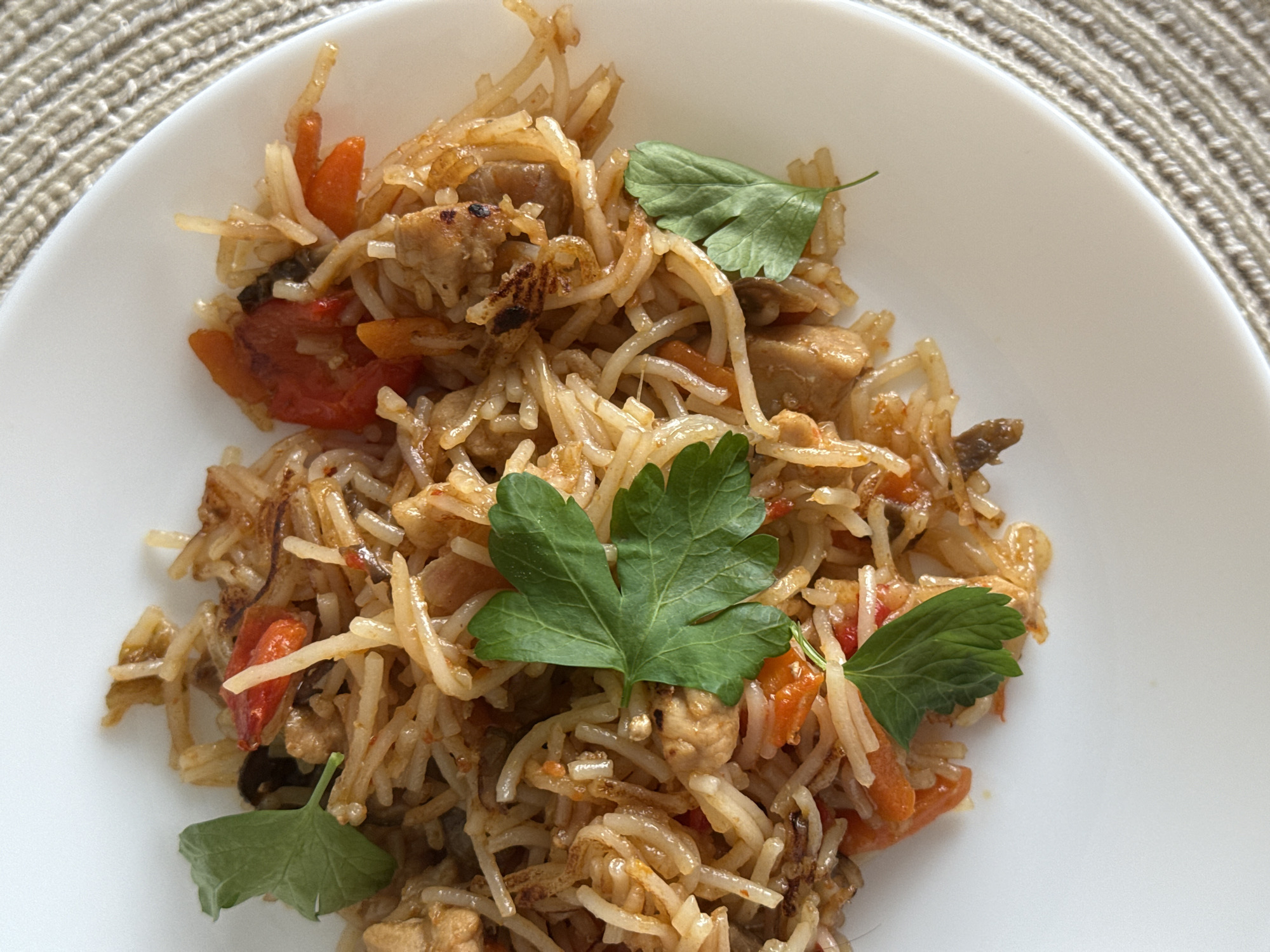A close-up, overhead shot features a portion of a white plate filled with a portion of a rice dish. The rice appears to be mixed with small pieces of meat, carrots, and tomato, and is garnished with a few sprigs of fresh parsley. The dish is presented on a textured, beige placemat. The specific location in the city of Гамово, Russia, cannot be determined from the visual information provided.