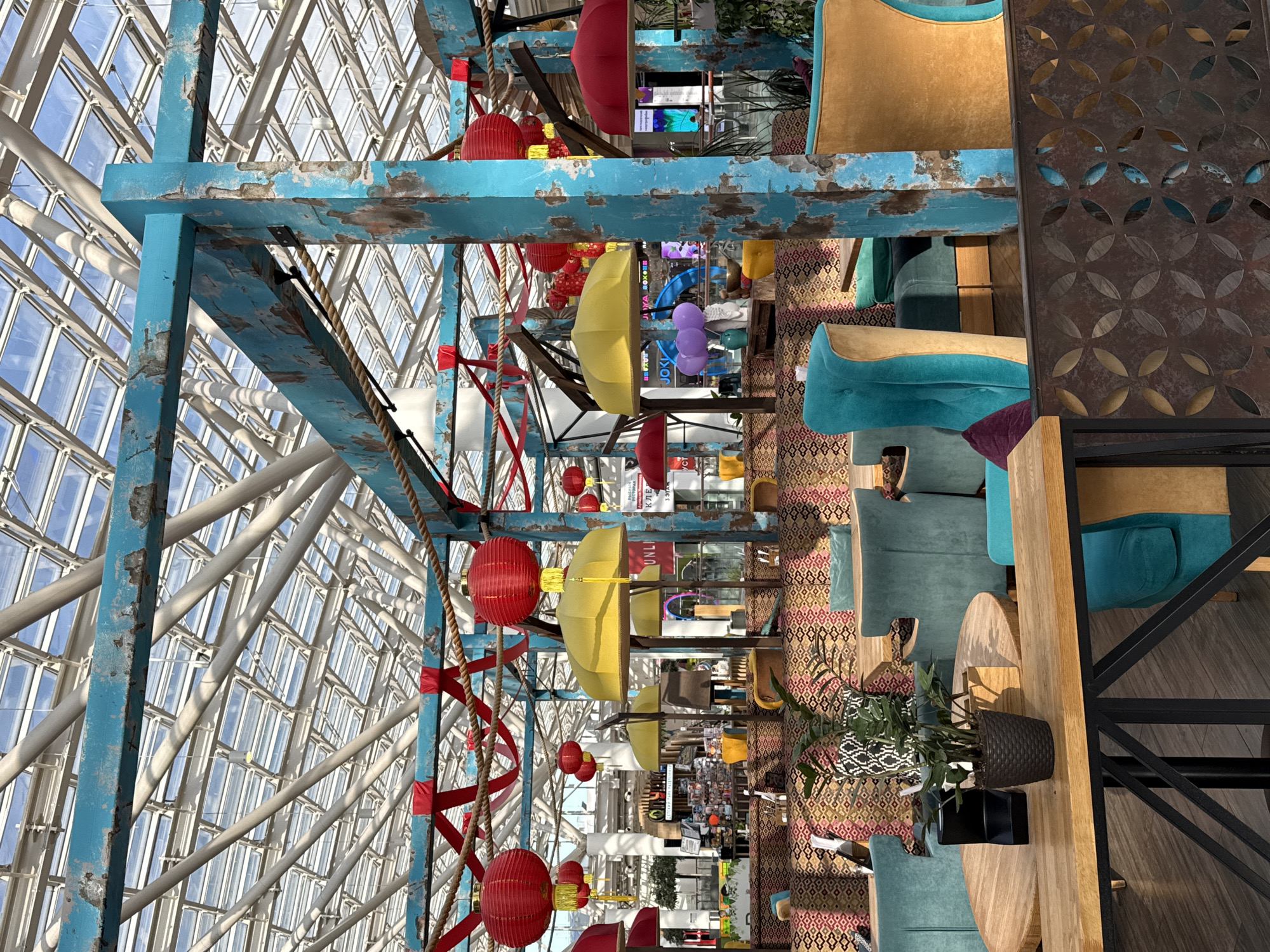 The image depicts an interior space in Khimki, Russia, presumably a restaurant or cafe. The architectural structure is prominent, featuring a high, geometrically patterned glass ceiling supported by a turquoise-painted metal frame. Decorations include red paper lanterns, yellow umbrellas, and red spiraling ribbons suspended from the framework. The space is furnished with turquoise and gold upholstered chairs, patterned rugs, and wooden tables adorned with potted plants. In the background, the image captures glimpses of other spaces and activities, with a combination of retail and recreational elements, including signage and displays, are visible.