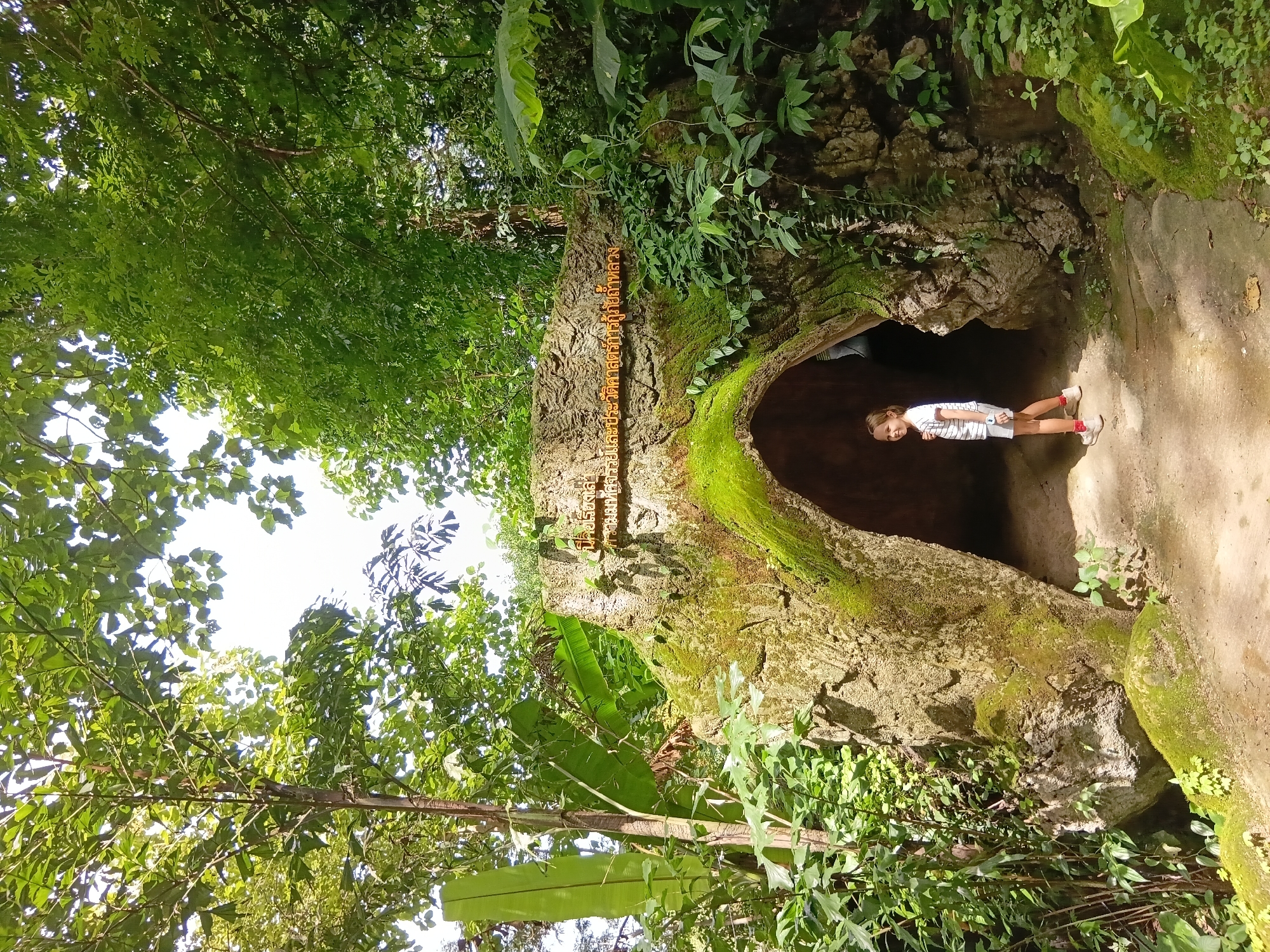 A young girl with blonde hair stands on a dirt path in front of a moss-covered cave entrance. She is wearing a striped t-shirt and shorts. The cave is surrounded by lush green foliage and trees, suggesting a natural outdoor setting. A sign with Thai script is visible on the rock above the cave entrance. The scene appears to be at Doi Nang Non, Mae Sai District, Thailand, a well-known natural attraction.