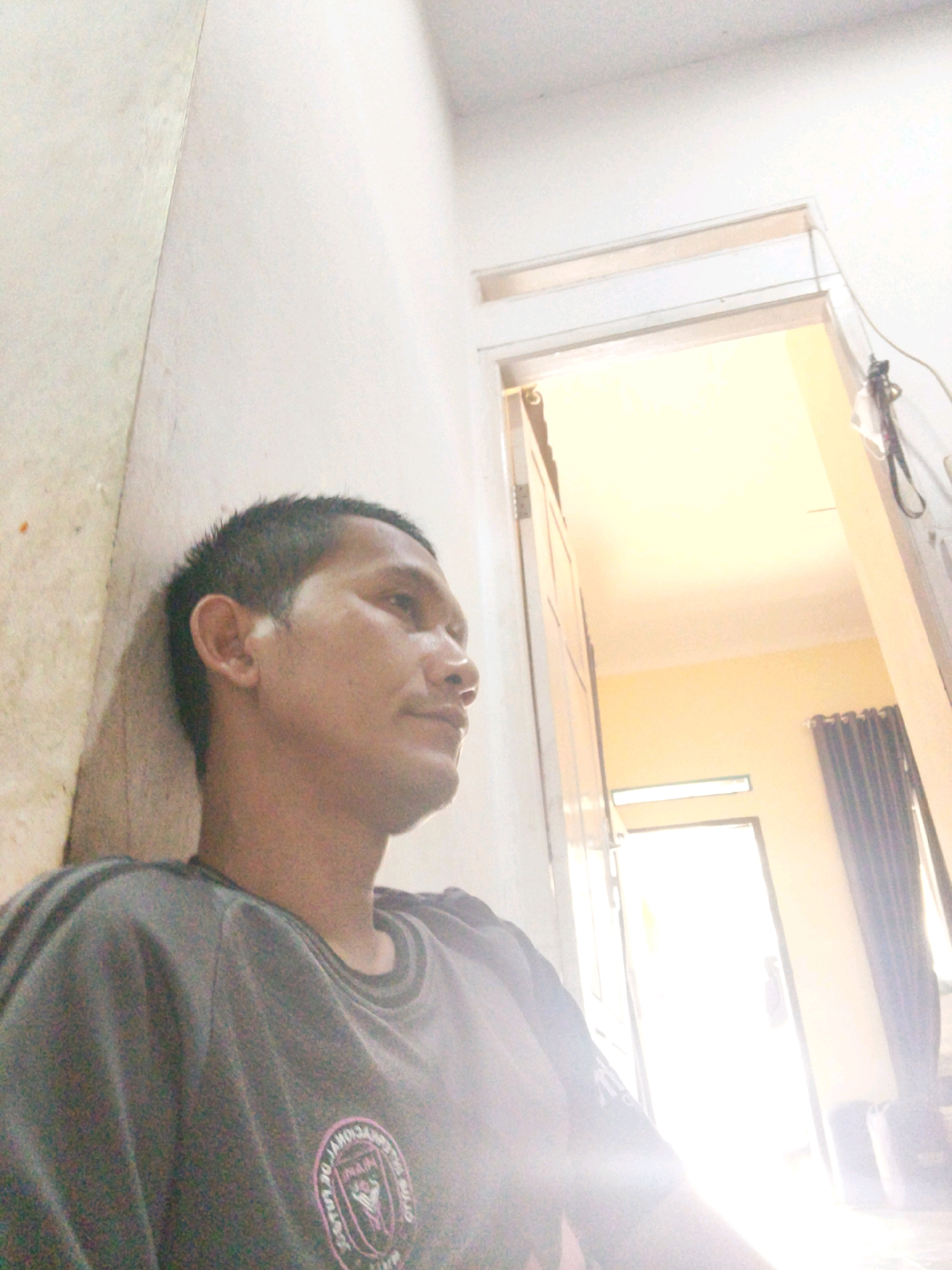The image is a selfie taken by a man leaning against a light-colored wall. He has short black hair and is wearing a dark gray t-shirt with a pink circular logo on the left side of his chest. The man is looking to his right, away from the camera. The background shows a doorway leading into a room with bright light, suggesting it's daytime or there is strong artificial lighting. A dark curtain is visible on the right side of the doorway. The location is identified as Banjar Agung, Indonesia.