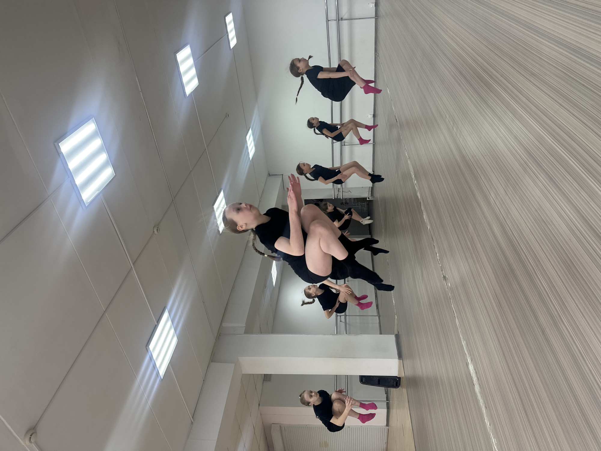 The image shows a dance studio in Votkinsk, Russia. Seven young girls in black leotards and pink or black socks are practicing a floor exercise. The studio has a mirrored wall and a barre. The floor is wood-patterned. The ceiling is white with rectangular fluorescent lights.