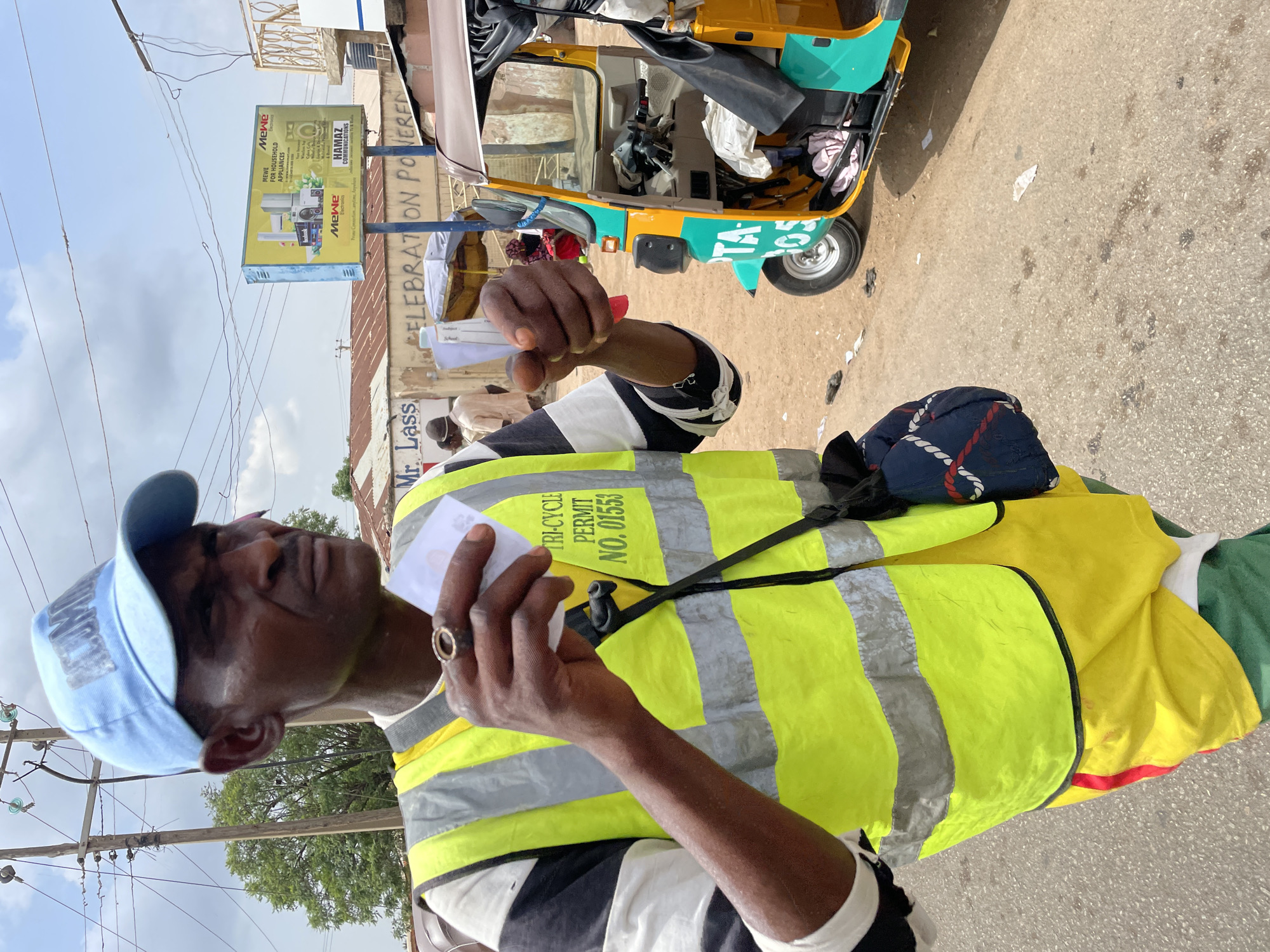 The image shows a man in a high-visibility yellow safety vest that reads "TRI-CYCLE PERMIT NO. 01553," holding a piece of paper. He is also wearing a blue cap. A tricycle (also known as a Keke Napep in Nigeria) is partially visible in the background. Street advertising signs are visible in the background and read "HAMAZ COMMUNICATIONS." Another sign mentions "MEWE" for household appliances. The letters "TA" are shown on the side of the tri-cycle. The surrounding environment suggests an urban setting in Jos, Nigeria.