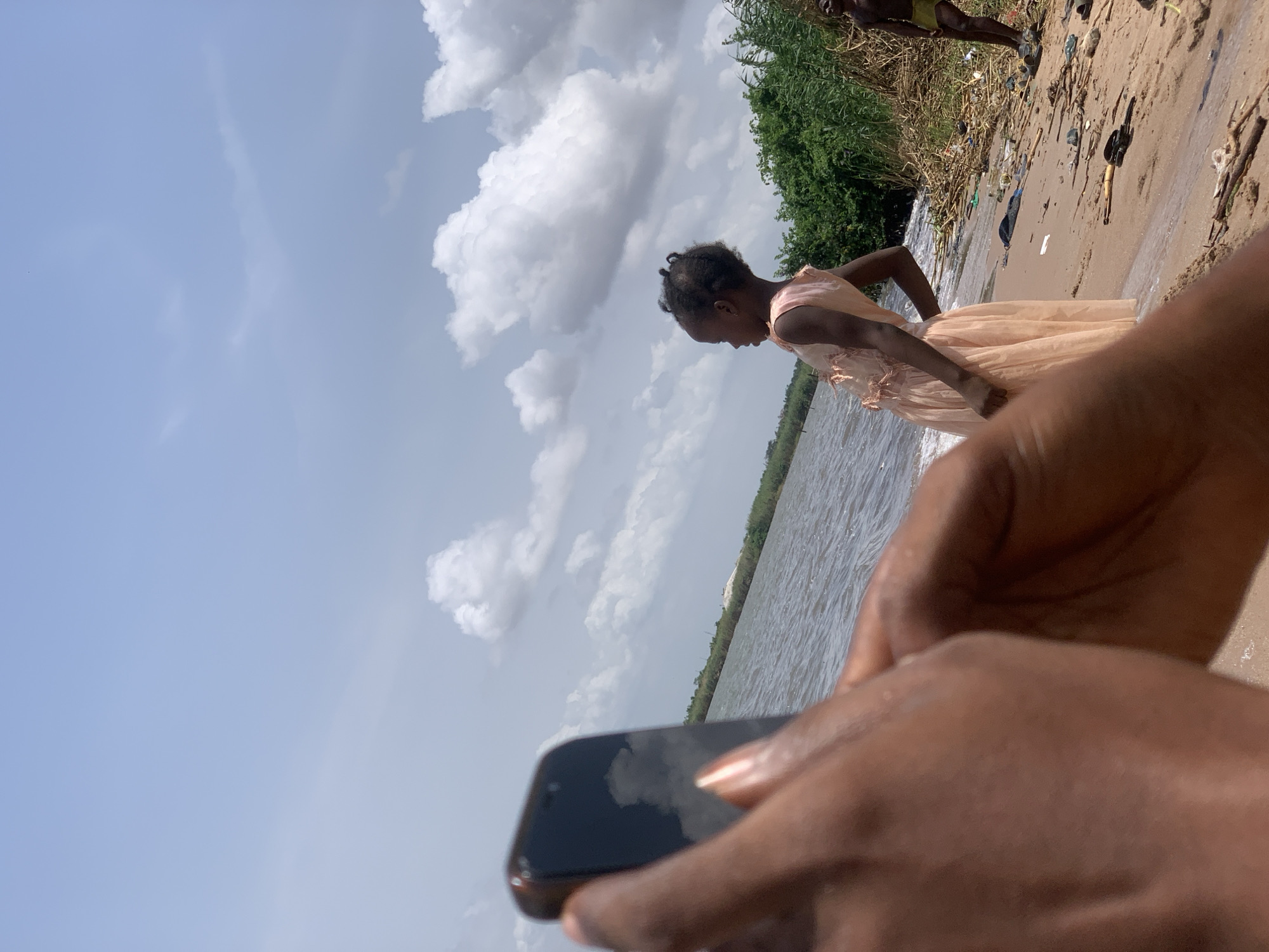A person is holding a black smartphone while standing on a sandy beach in Ikorodu, Nigeria. The phone's screen reflects the sky and clouds. A young girl in a light-colored dress stands near the water's edge. In the background, there is vegetation and a body of water. Debris litters the shoreline in the upper right of the frame. Another person's legs are visible further in the background.