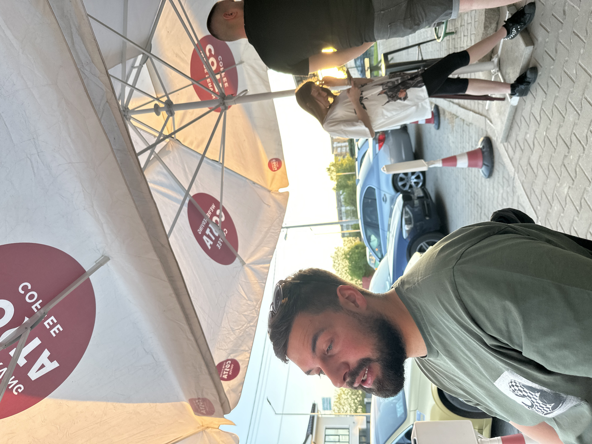 The image shows an outdoor scene in Plovdiv, Bulgaria. A man with a beard wearing a green shirt is in the foreground, looking down. Behind him is an open umbrella with the "Costa Coffee" logo on it. In the background, a woman wearing a white shirt and black pants is standing with a man wearing a black shirt and gray shorts. There is a safety cone next to the woman. Cars are parked behind them. The ground appears to be paved with interlocking pavers.