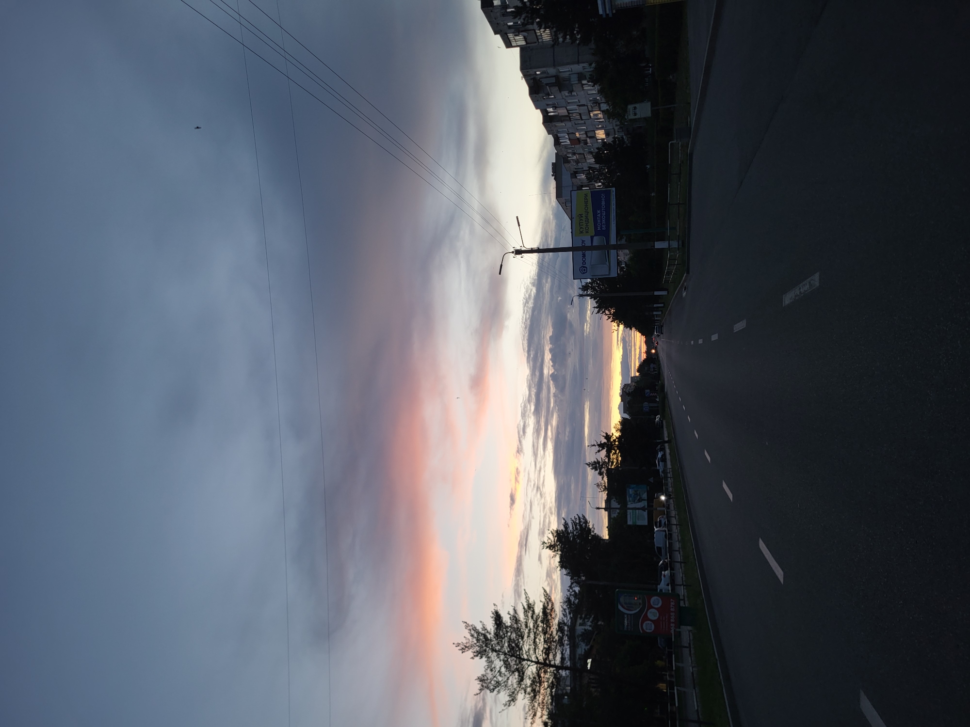 The image depicts an asphalt road in Horishni Plavni, Ukraine, at dusk. The sky is filled with dramatic clouds, with hues of pink, orange, and gray. A flock of birds is visible in the sky, along with power lines crisscrossing overhead. On the right side of the road, several apartment buildings are visible, their windows reflecting the fading light. Trees line the left side of the road, interspersed with streetlights and billboards. Cars are present on the road, though their details are obscured by the low light conditions. The overall scene is one of a calm, urban evening setting as day transitions into night.