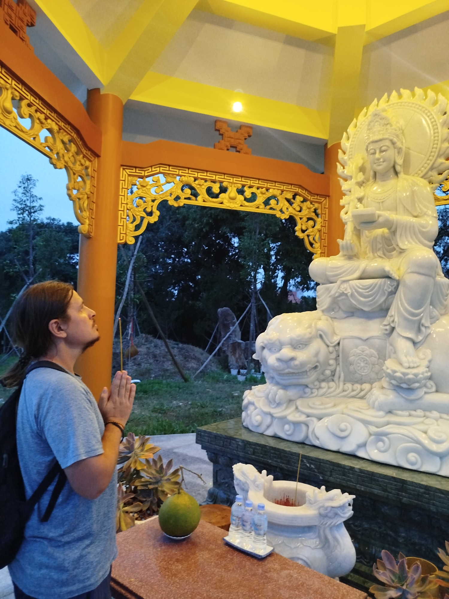 An outdoor scene captures a man in a state of reverence at a Buddhist shrine, likely in Phú An, Vietnam, during dusk or early evening. The man, positioned on the left side of the frame and viewed in profile, has dark hair tied in a ponytail and is wearing a grey t-shirt and a dark backpack. His hands are clasped in a prayer gesture, holding a burning incense stick, as he looks upwards towards a large, serene white Buddha or Bodhisattva statue.
The statue, made of white marble, depicts a figure seated on a lotus base, which in turn rests upon a white, foo dog or lion-like creature. A radiant halo-like mandorla is visible behind the statue's head. The shrine's structure features ornate gold carvings along its borders, bright orange columns, and a vivid yellow ceiling, which is illuminated.
In the foreground, a brownish-red altar holds offerings including a large green, round fruit (possibly a pomelo), a white ceramic incense burner adorned with dragon motifs and containing burning incense sticks, and a small tray holding three plastic water bottles. Succulent plants are arranged near the altar, and lush greenery, including trees, fills the background under a muted blue sky, indicating the outdoor setting. The visible incense smoke and the man's posture convey an atmosphere of devotion and tranquility. No discernible text is visible in the image.