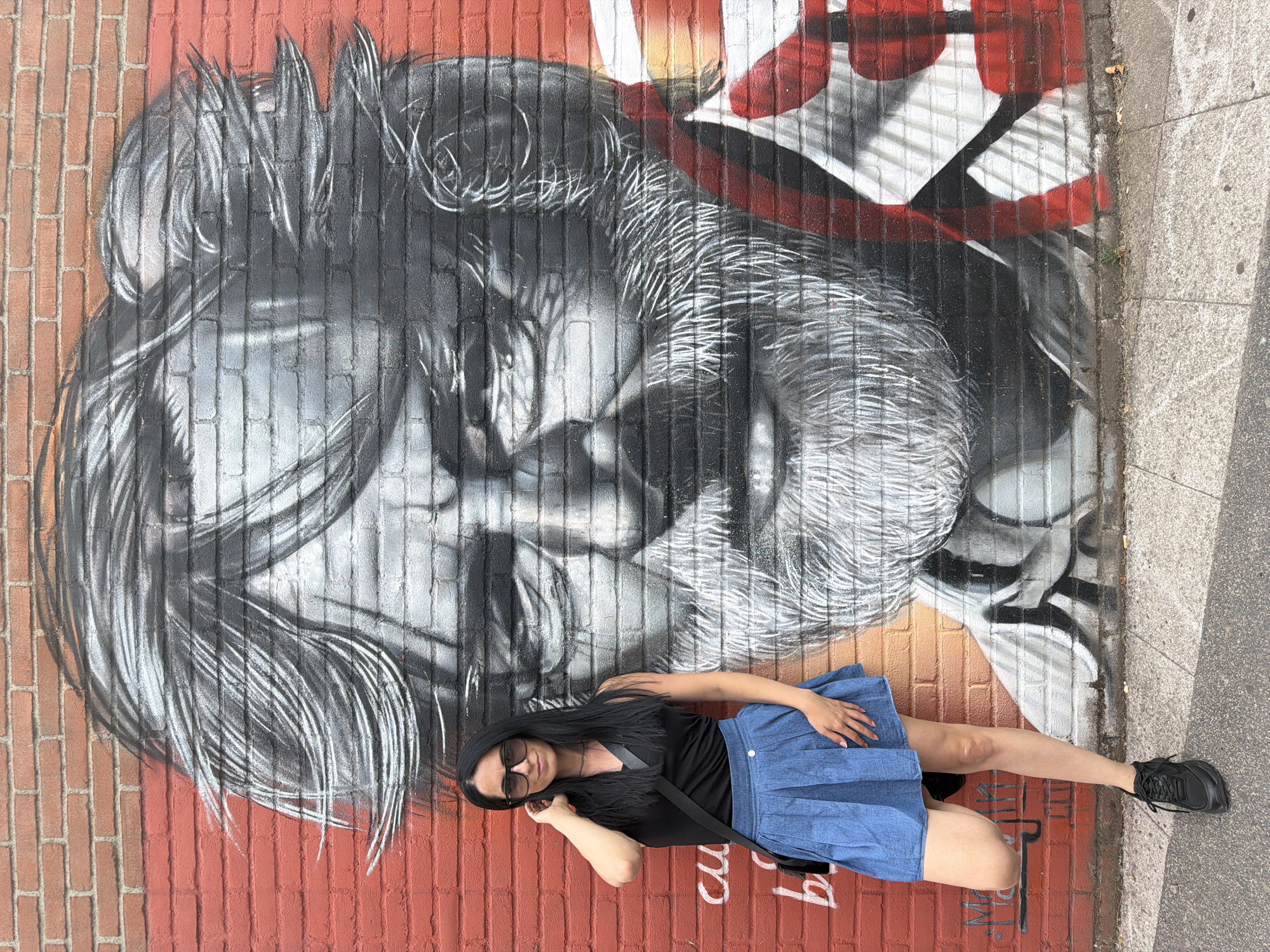 A woman with long, dark hair and sunglasses stands in front of a brick wall adorned with a large mural of a man's face. The mural is rendered in shades of black, white, and gray, with hints of red and orange. The woman is wearing a black sleeveless top, a blue pleated skirt, and black sneakers. She has one leg bent and leaning against the wall, and her right hand is raised to her hair. A black crossbody bag is slung over her left shoulder. The brick wall is red and textured. The ground in front of the wall is a sidewalk made of concrete tiles. The scene appears to be outdoors, possibly on a street in the city of Corsico, Italy.