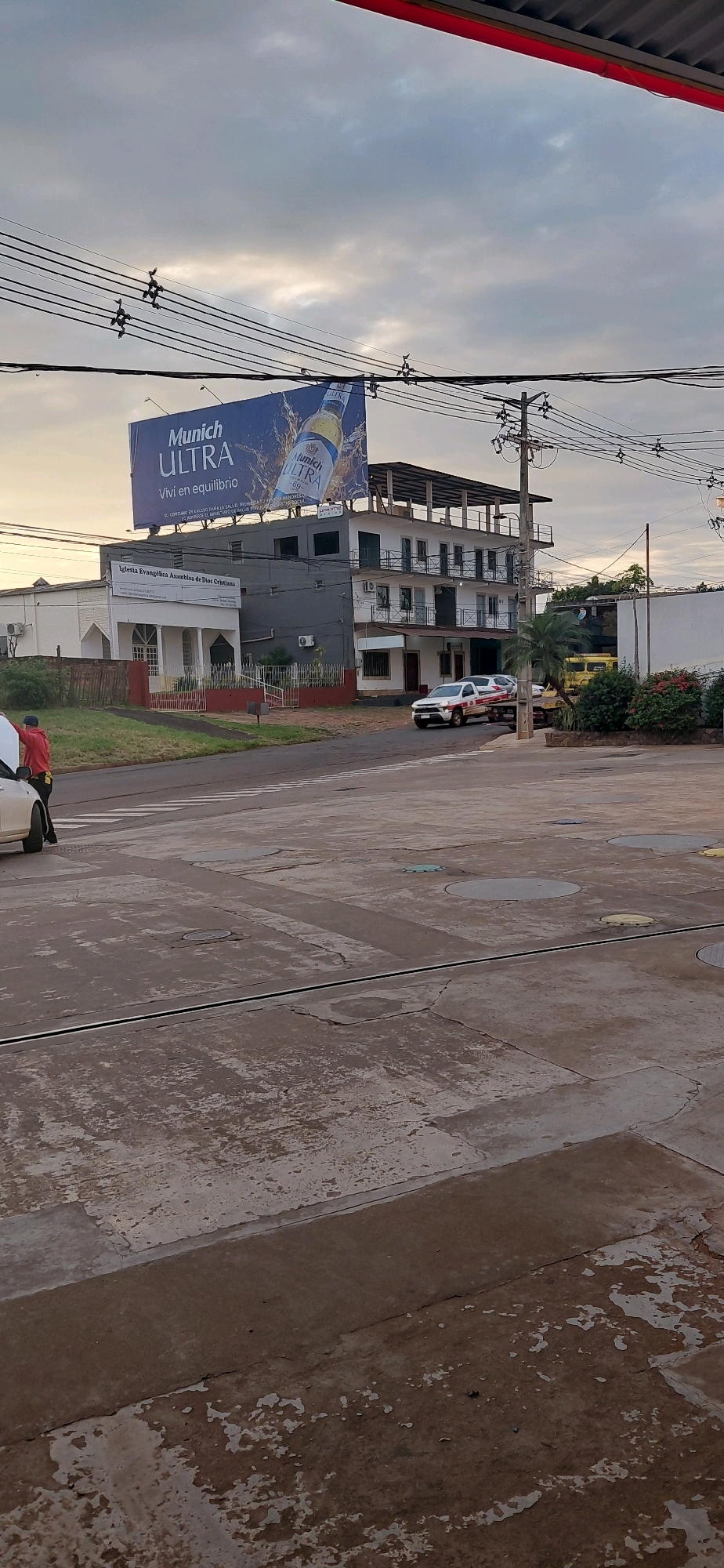 The image shows a street scene in Encarnación, Paraguay, with a multi-story building and a billboard. The billboard advertises "Munich ULTRA" beer. A white utility vehicle with red accents is parked on the right side of the street, facing left. In the foreground, a person wearing a red jacket and dark pants is standing next to a white car. The street is paved and shows signs of wear, with cracks and stains. The sky is cloudy. There are power lines running across the top of the image, with several birds perched on them. A church building, labeled "Iglesia Evangélica Asamble de Dios Cristiana," is visible to the left of the multi-story building.