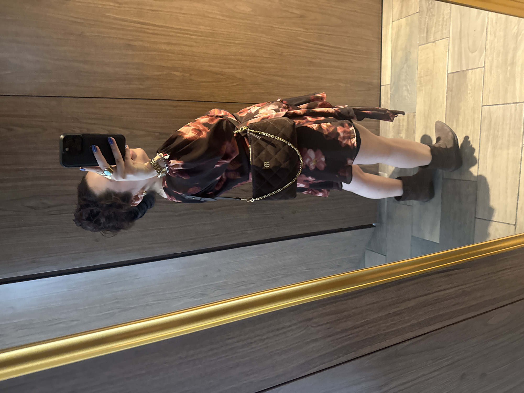 The image shows a woman taking a selfie in front of a mirror. The location is unclear, but it is likely in Mexico City, Mexico. She has dark hair and is wearing a floral dress, brown ankle boots, and a black quilted purse with a gold chain. She is holding a black smartphone with blue fingernails. The background includes wood paneling and tiled flooring. The mirror is framed with a gold border.