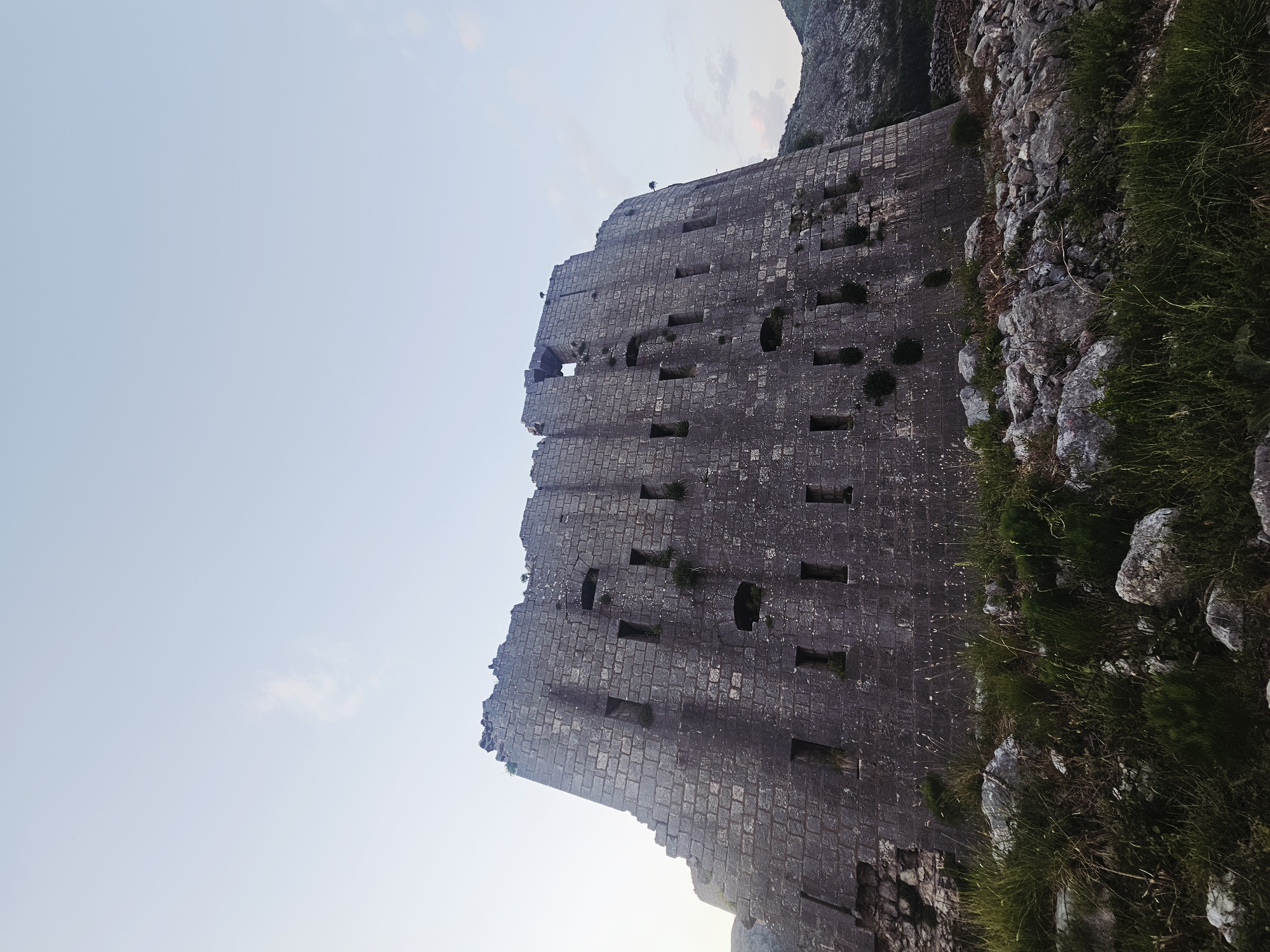 Here is a description of the image:
The image depicts the ruins of St John's Fortress located in Kotor, Montenegro. The fortress is constructed from grey stone blocks and exhibits numerous rectangular openings along its walls. The structure is situated adjacent to a steep, rocky terrain covered with grass and vegetation. The sky is visible behind the fortress, displaying a light blue gradient.
