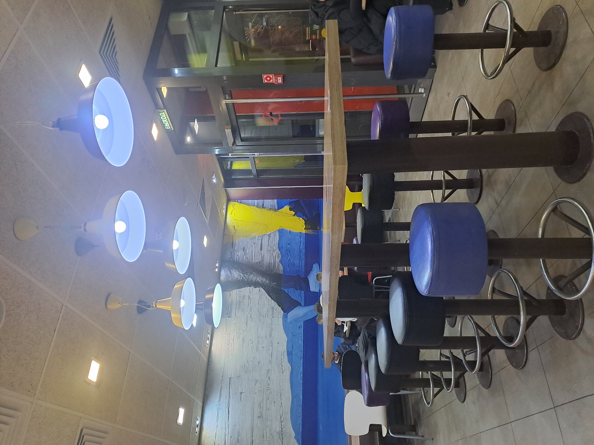 The image shows the interior of a McDonald's restaurant in Elektrostal, Russia. The scene includes rows of high-top tables with attached stools, some upholstered in blue, purple, or black. People are seated at the tables, dining. The walls are decorated with a mural featuring blue and yellow colors. A row of pendant lamps with wide, dish-shaped shades illuminates the area. A glass-enclosed section is visible in the background. A "No Smoking" sign is displayed.