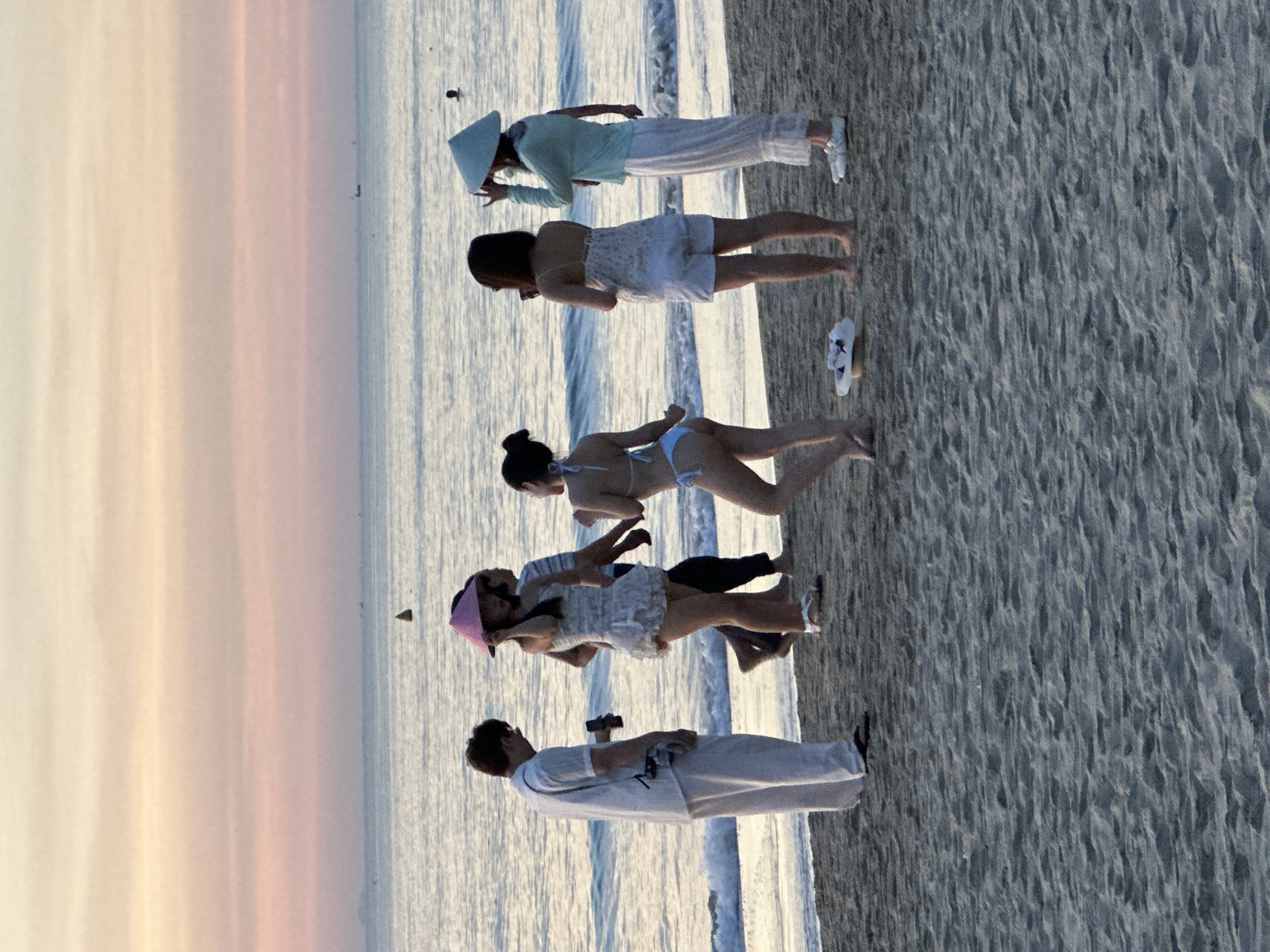 The image captures a scene on a beach in Đà Nẵng, Vietnam, during what appears to be sunset given the soft, pastel hues of the sky. Several individuals are present on the sand near the water's edge.
On the left, a man in a white long-sleeved shirt and loose white pants stands facing away from the camera, holding a dark object, possibly a phone, in his right hand. He is wearing dark flip-flops.
Moving towards the center, a woman in a short, white, ruffled dress and sandals is visible, her body turned to the side. She appears to be looking at something in the distance. Behind her and to her right, another person, possibly a woman, is visible wearing a striped shirt.
Further to the right, a woman in a light blue long-sleeved top and white pants, wearing a conical hat, stands with her right hand raised near her head, as if adjusting the hat or gesturing. She is also wearing white shoes. To her left, a woman in a white bikini is seen from the back, her body in motion as if dancing or walking.
In the foreground on the sand, a white hat with some dark objects on it is placed near the bikini-clad woman. The beach is covered in sand, with gentle waves lapping at the shore. The ocean stretches out to the horizon, under a sky painted with soft oranges, pinks, and yellows. A few small dark shapes, possibly buoys or distant boats, are visible on the water. The overall atmosphere is relaxed and casual, with the figures enjoying the beach and the sunset.