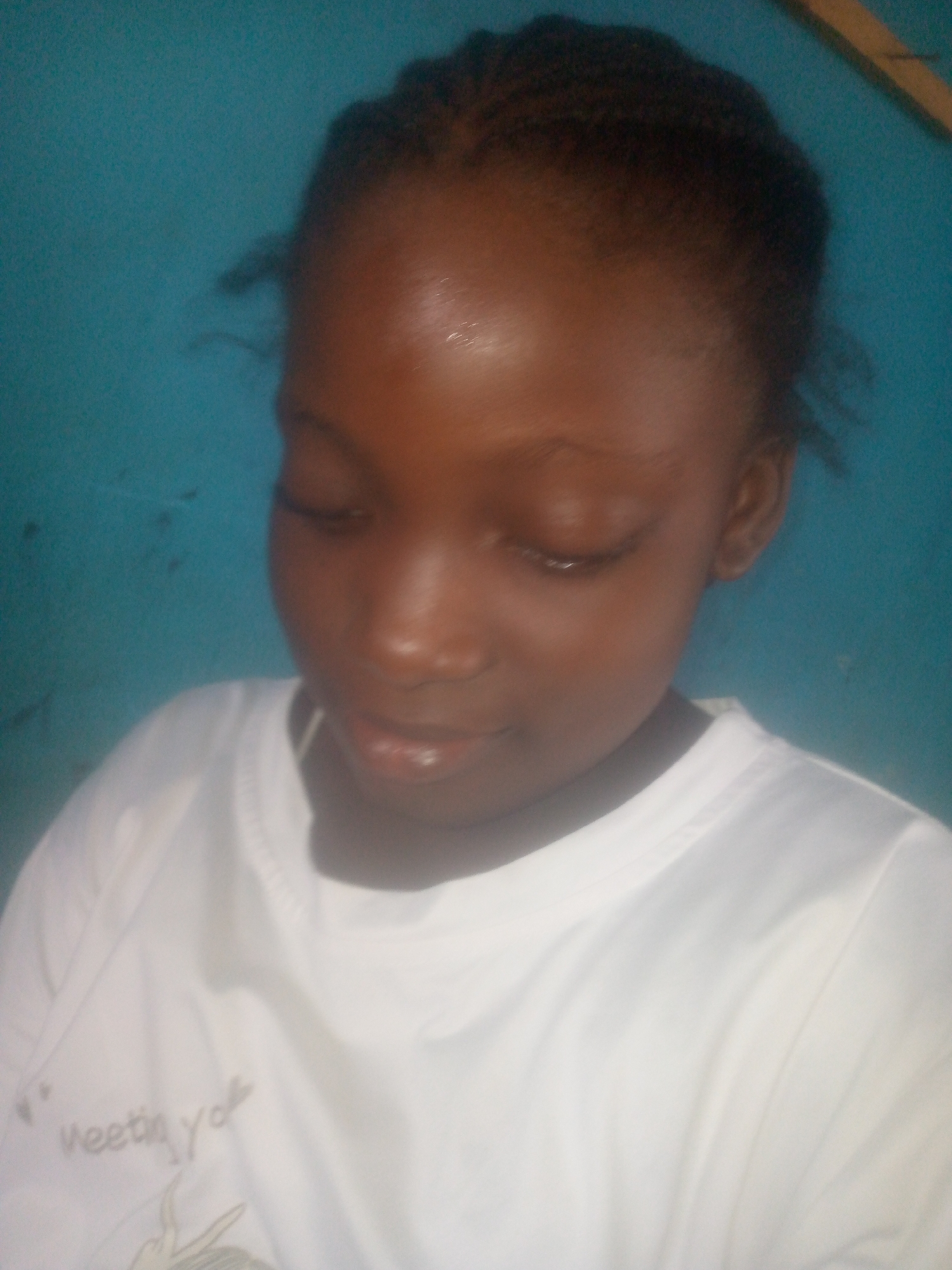 A young, dark-skinned girl with braided hair is looking downwards. She is wearing a white t-shirt. The background is a plain blue wall. The location, Ikorodu, Nigeria, is not evident from the image's visual content.