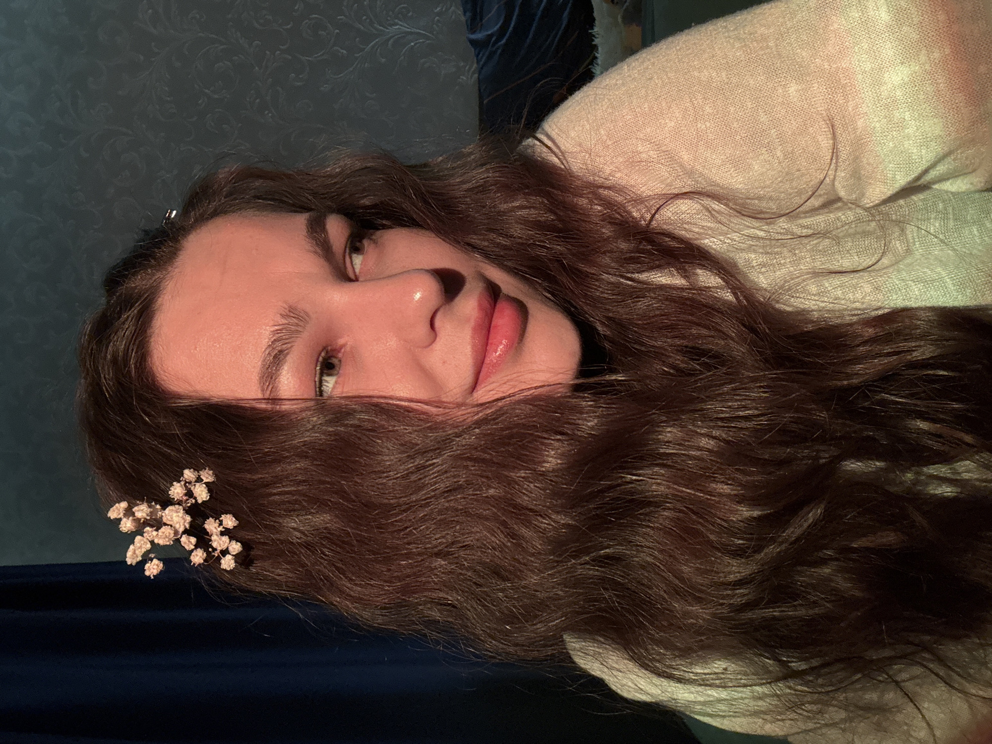 A close-up of a fair-skinned young woman with long, wavy brown hair and green eyes is presented at an angle. Her face is partially obscured by her hair, which is adorned with a sprig of small, white flowers near her forehead. The woman is wearing a light-colored sweater. The background is divided, with a patterned wall behind her head and a dark blue fabric below. There is no information provided to indicate the location is in Chaikovsky, Russia.
